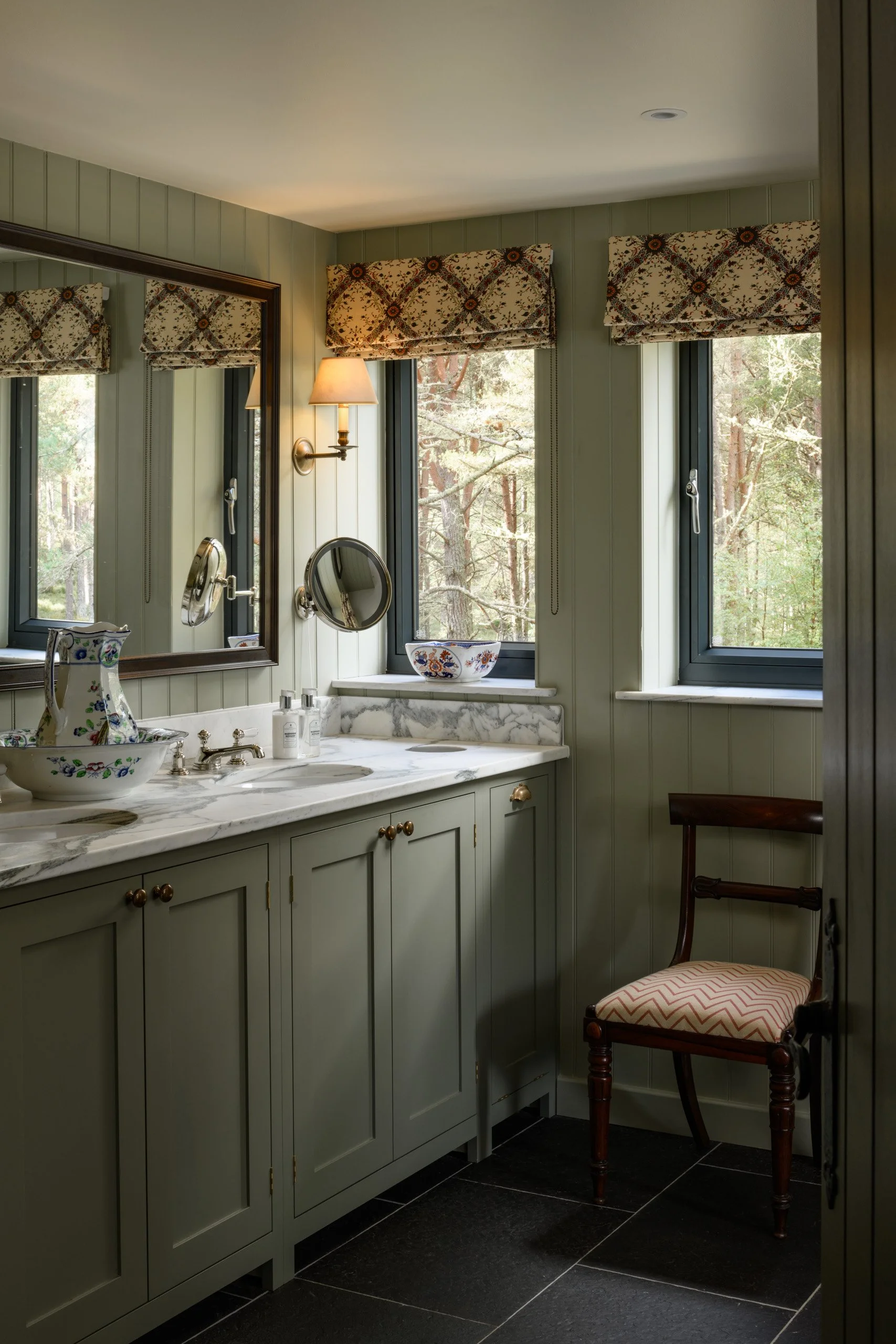 A cozy bathroom with a double marble sink vanity, a large mirror, a wall-mounted light, two windows with patterned valances, and a wooden chair with an upholstered seat.