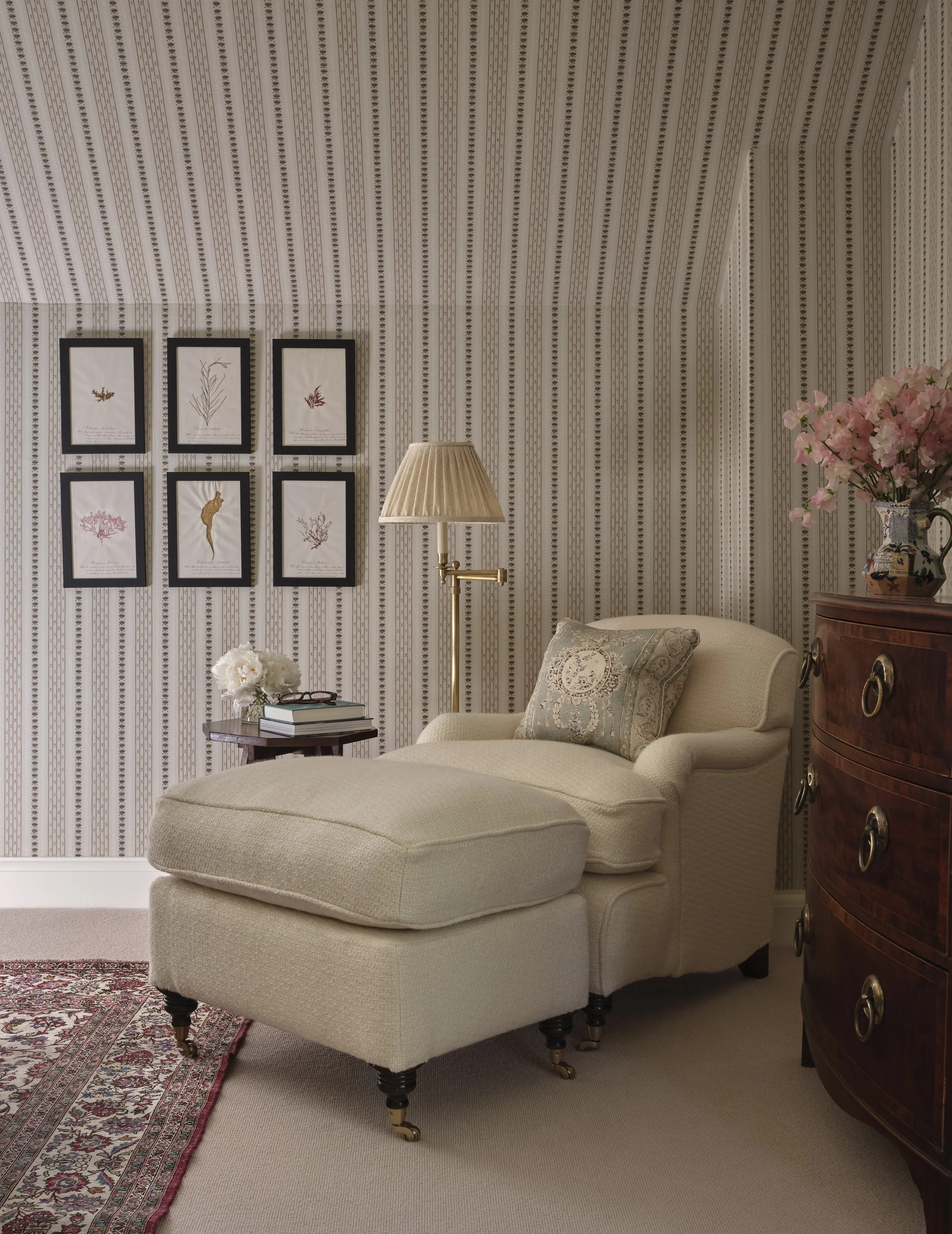 A cozy living room corner with a beige armchair and matching ottoman, a wooden side table with white flowers and books, a brass floor lamp with a pleated shade, a curved wooden dresser with a vase of pink flowers, patterned wallpaper, framed botanical art on the wall, and a red patterned rug on a cream carpeted floor.