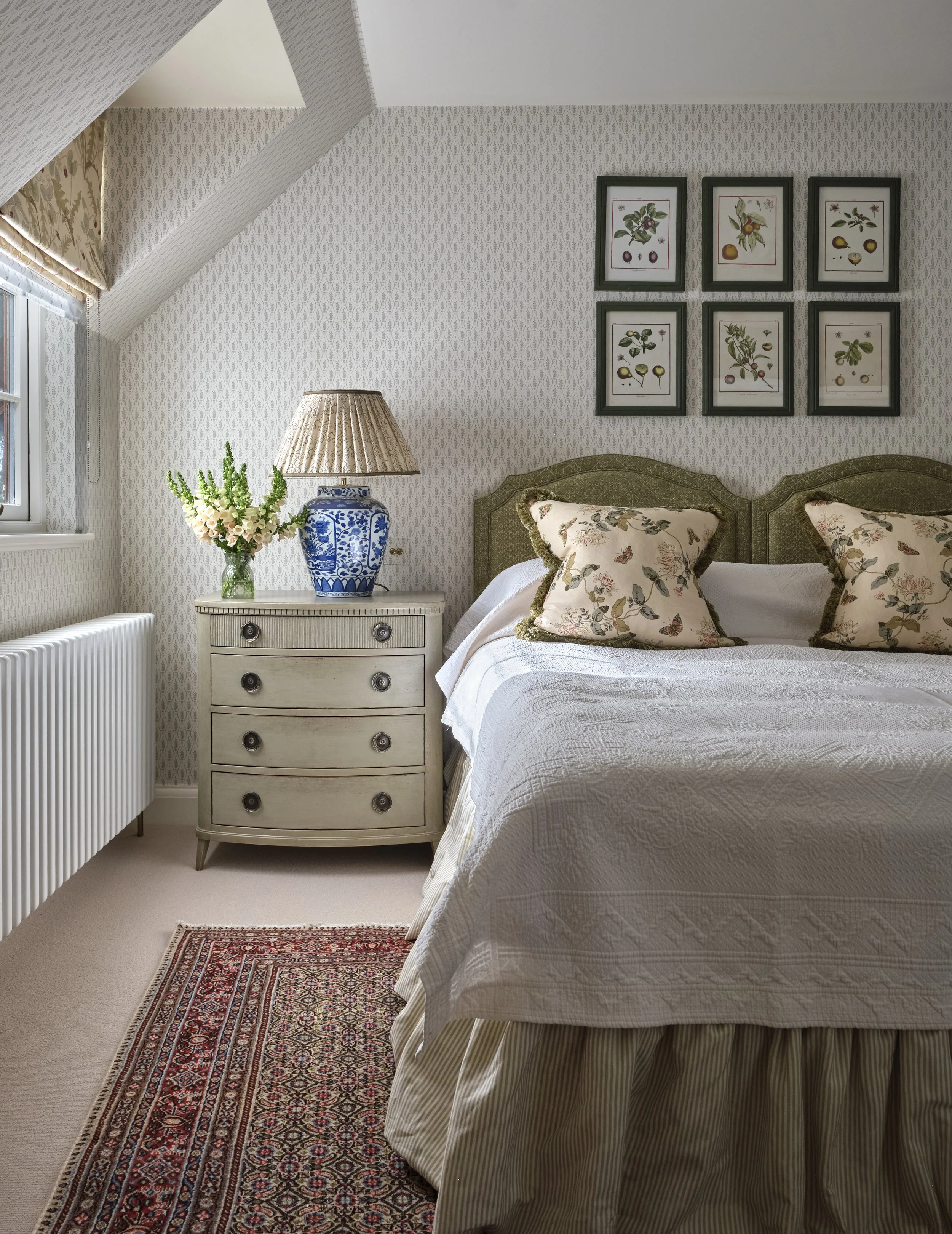 A cozy bedroom with a white bed, floral pillows, a vintage dresser with a blue and white lamp, a vase with flowers, framed botanical art on the wall, a window with a floral valance, a radiator, and a patterned rug.