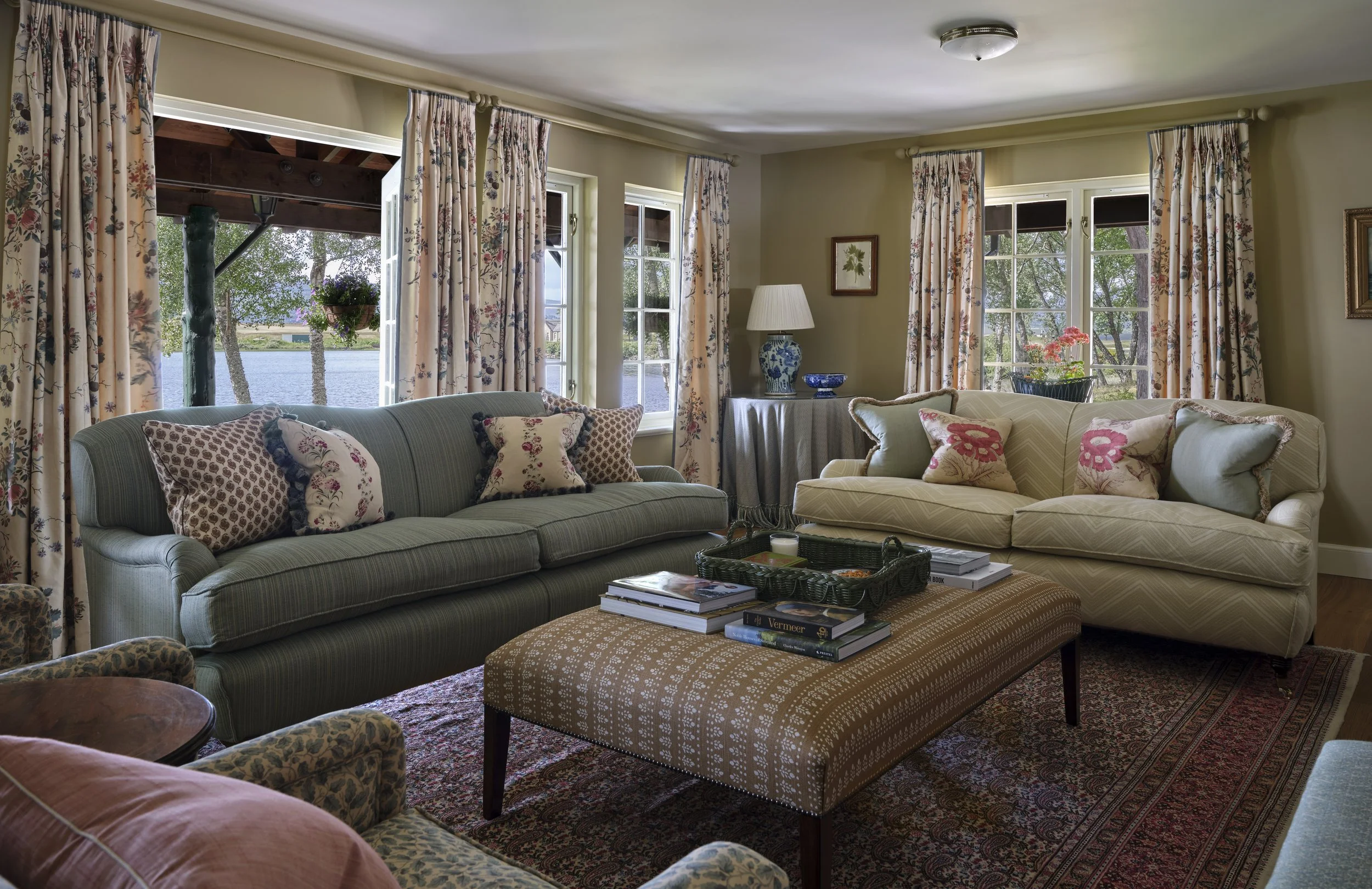 Living room with two sofas, an ottoman, a patterned rug, and windows overlooking a lake.