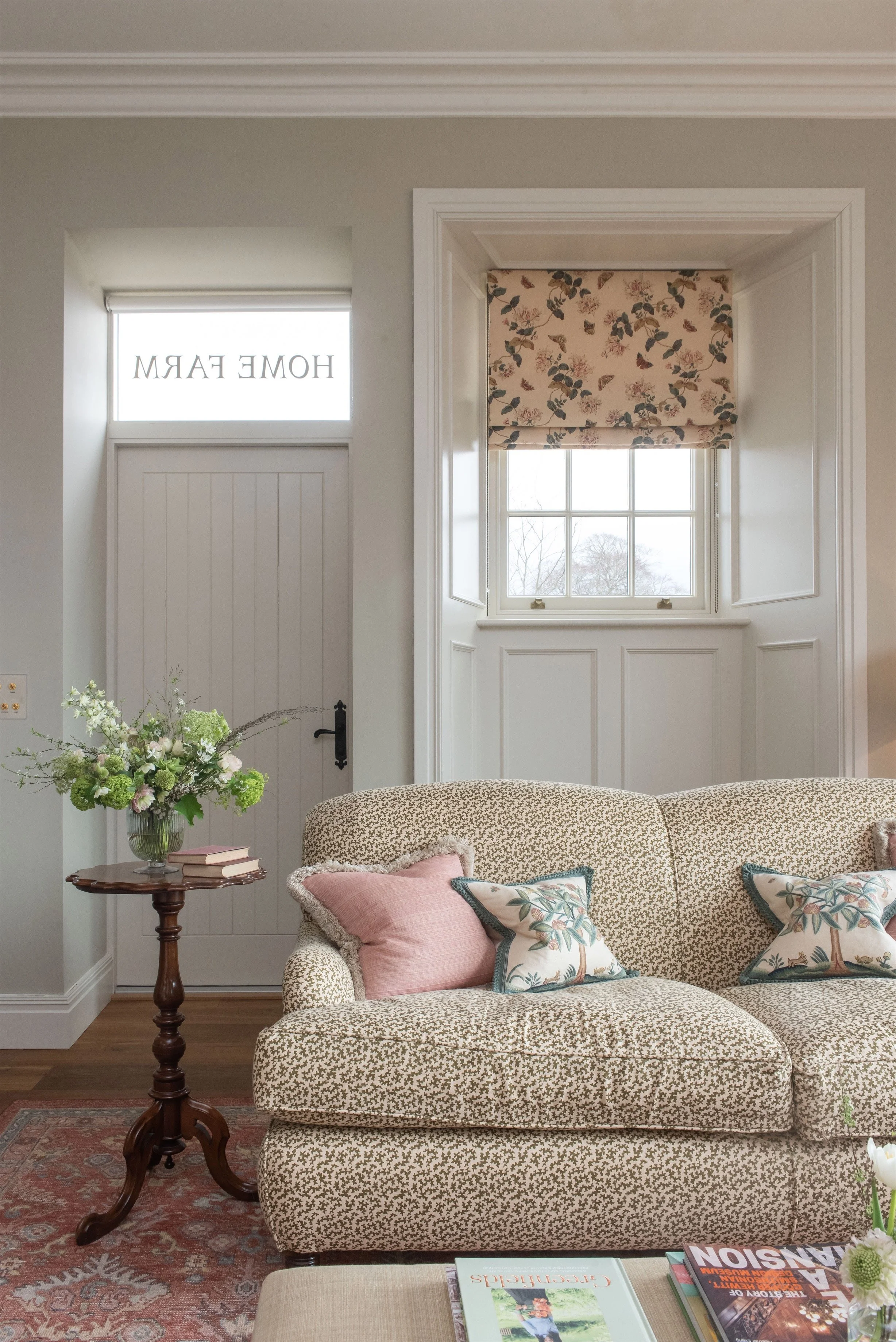 Cozy living room with a floral-patterned window shade, beige sofa with floral and pink pillows, a wooden side table with a vase of flowers and books, and a window with white trim. Decor includes a pink rug and a stack of magazines.