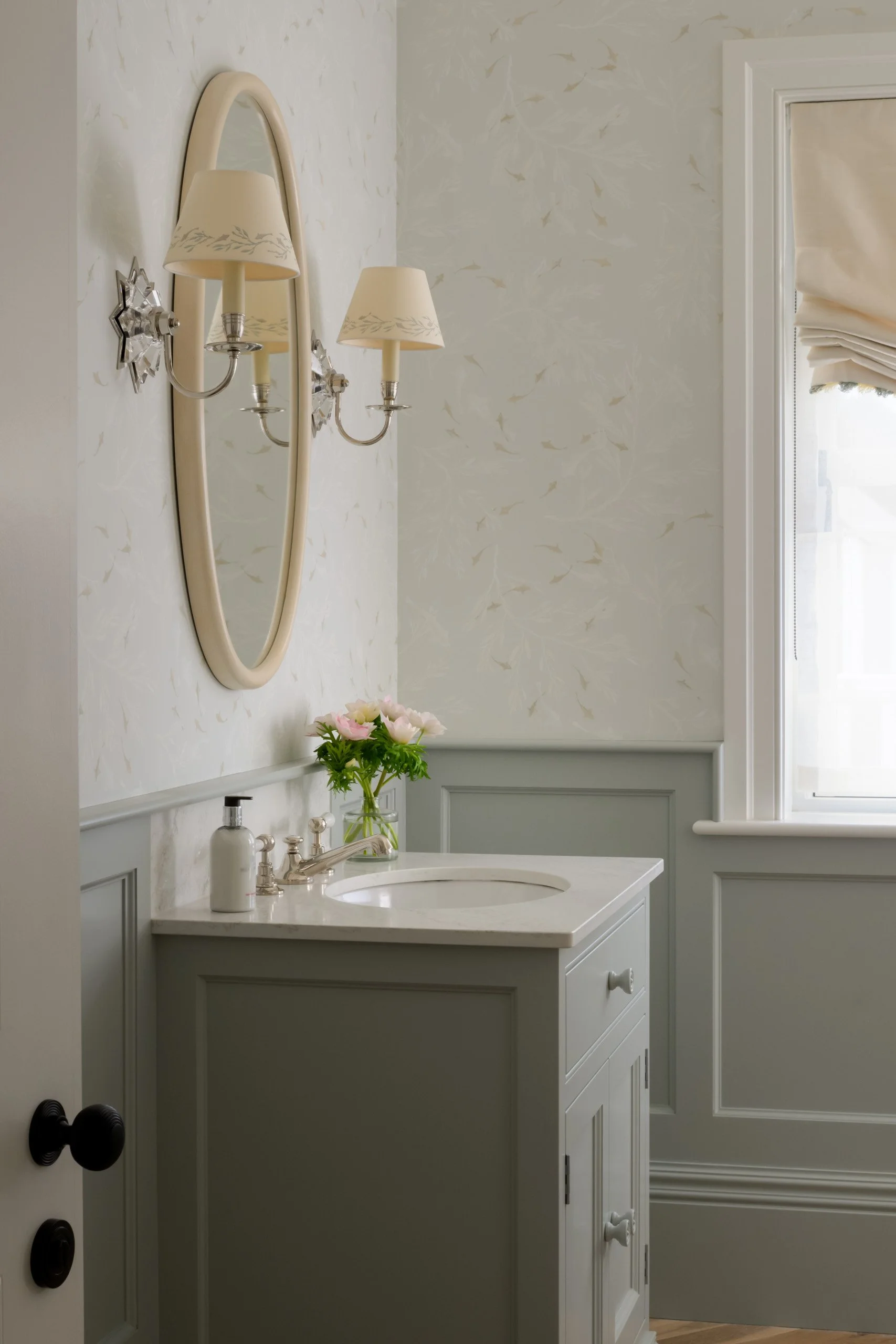 A bathroom with a light-colored vanity, a round mirror, a flower in a glass vase, wall-mounted lamps with cream shades, and a window with a beige window shade.