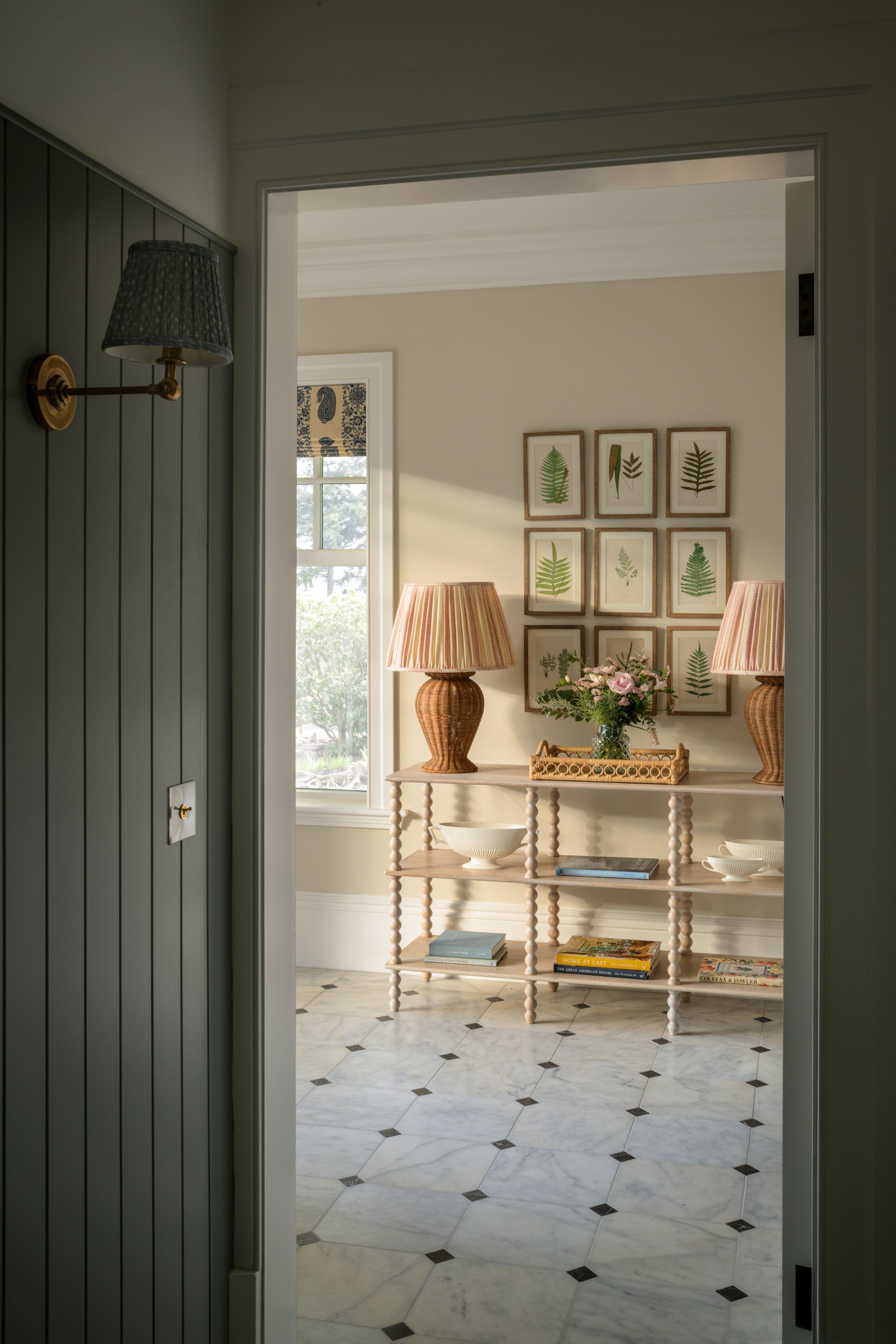 view through doorway into a corner of a room with a wooden console table, two woven lamps, a bouquet of pink flowers, framed leaf prints on the wall, and a marble floor with black diamond accents.