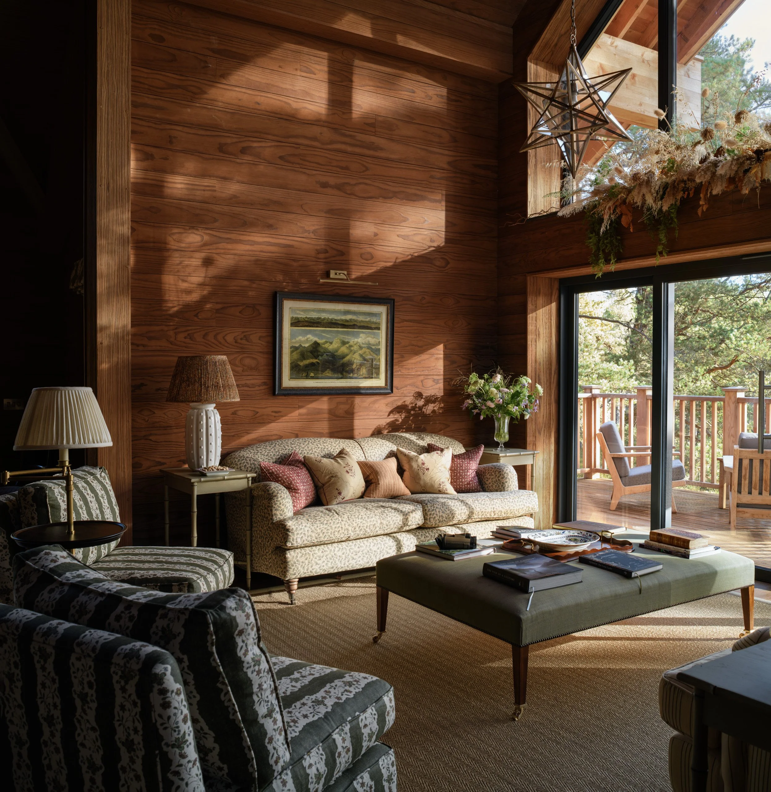 A cozy living room with wooden walls and large windows overlooking a balcony with outdoor furniture. The room has a patterned armchair, a sofa with throw pillows, a coffee table with books, and lamps, one on a side table and another on a stand, with 