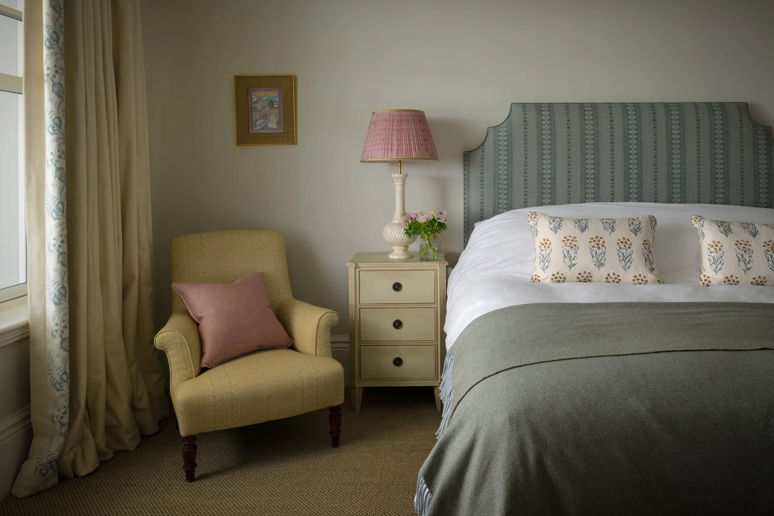A cozy bedroom featuring a bed with floral pillows, a lamp on a nightstand with pink flowers, a yellow armchair with a pink pillow by a window with curtains, and a framed picture on the wall.