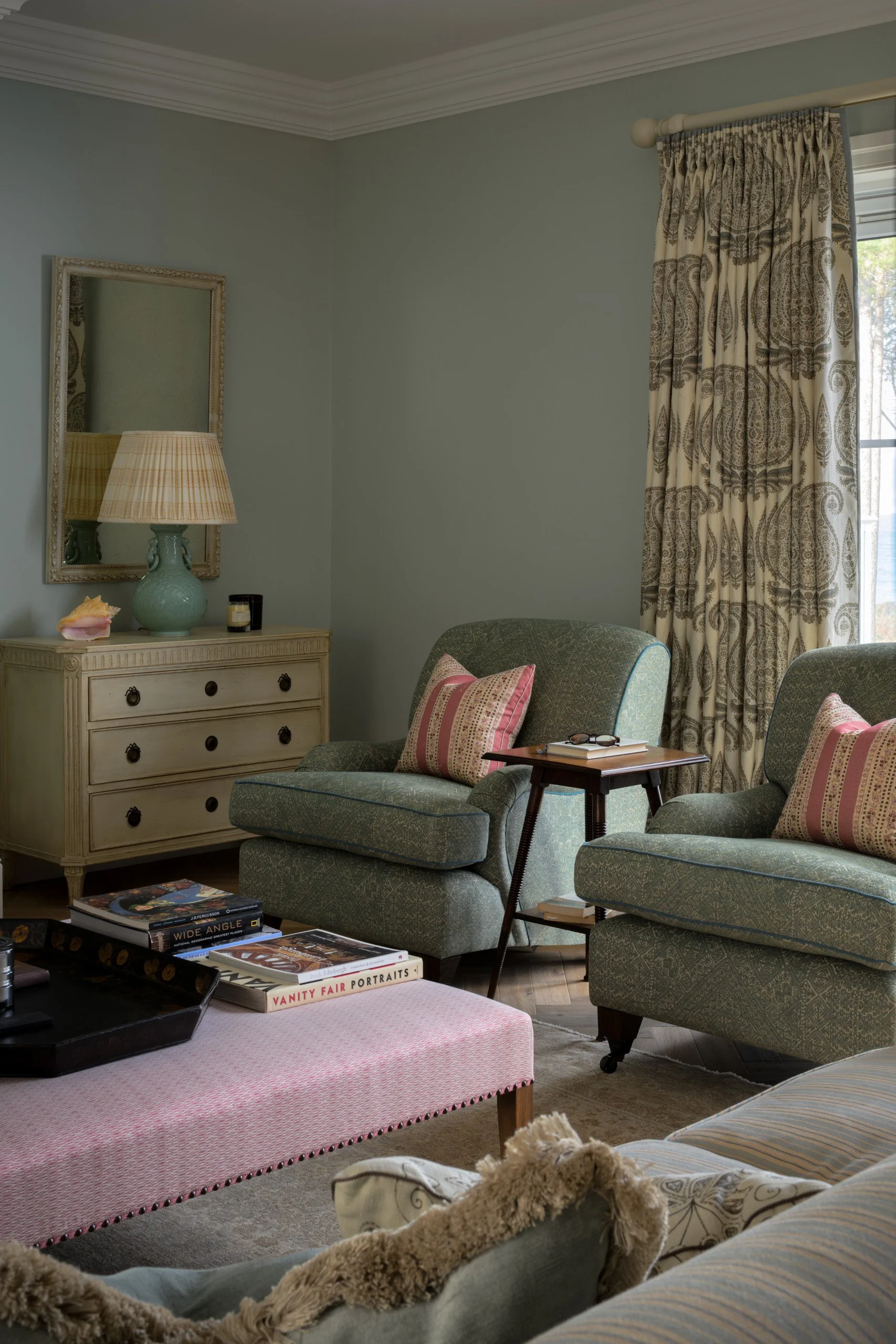 A cozy living room with two green armchairs, a pink upholstered bench, and a beige side table with books and a tray. There is a dresser with a mirror, a lamp, and candles. A window with patterned curtains lets in natural light.