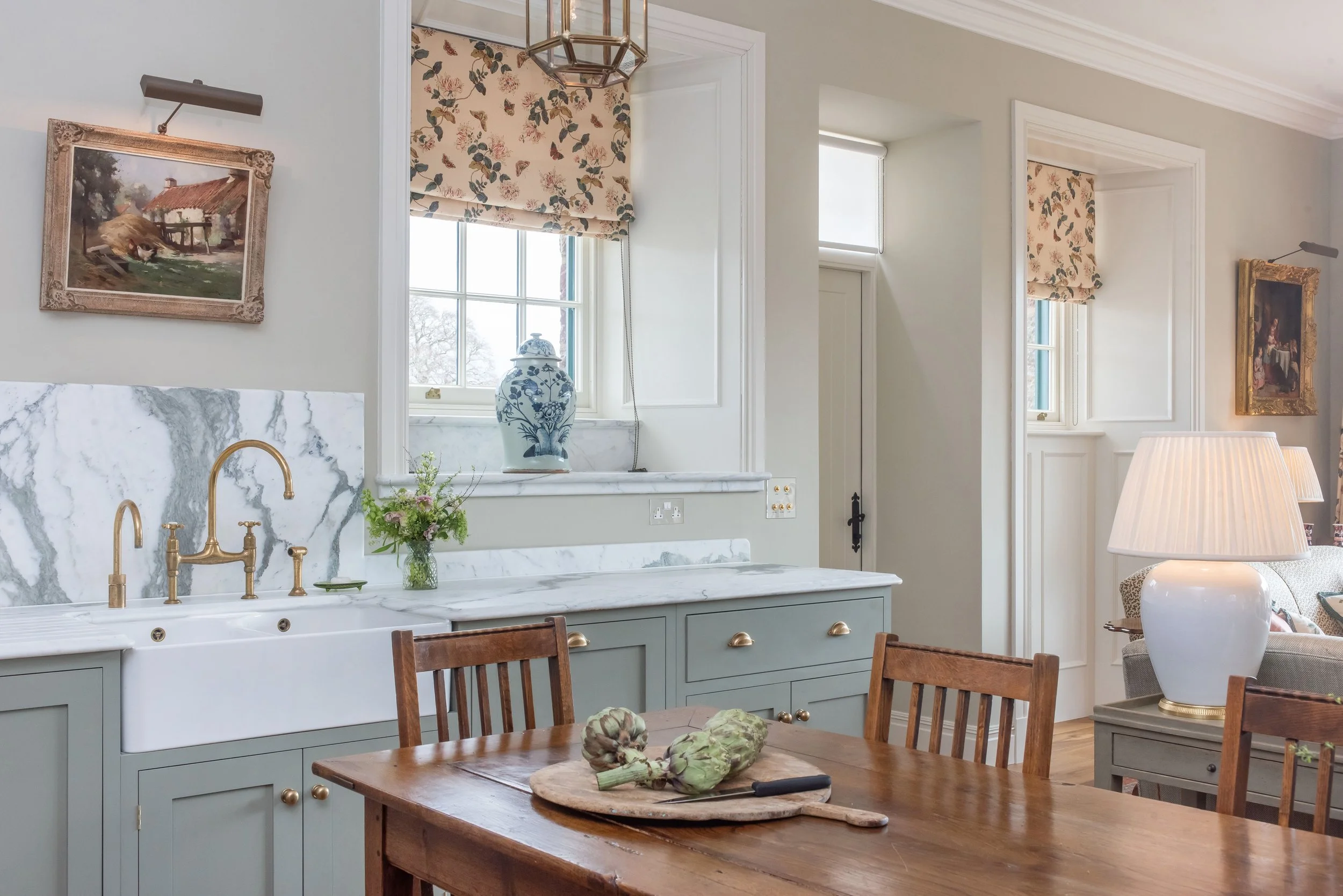 A kitchen with a wooden dining table, artichokes on a cutting board, a white farmhouse sink with brass faucet, window with floral Roman shade, and decorative vases, with light-colored walls and cabinetry.