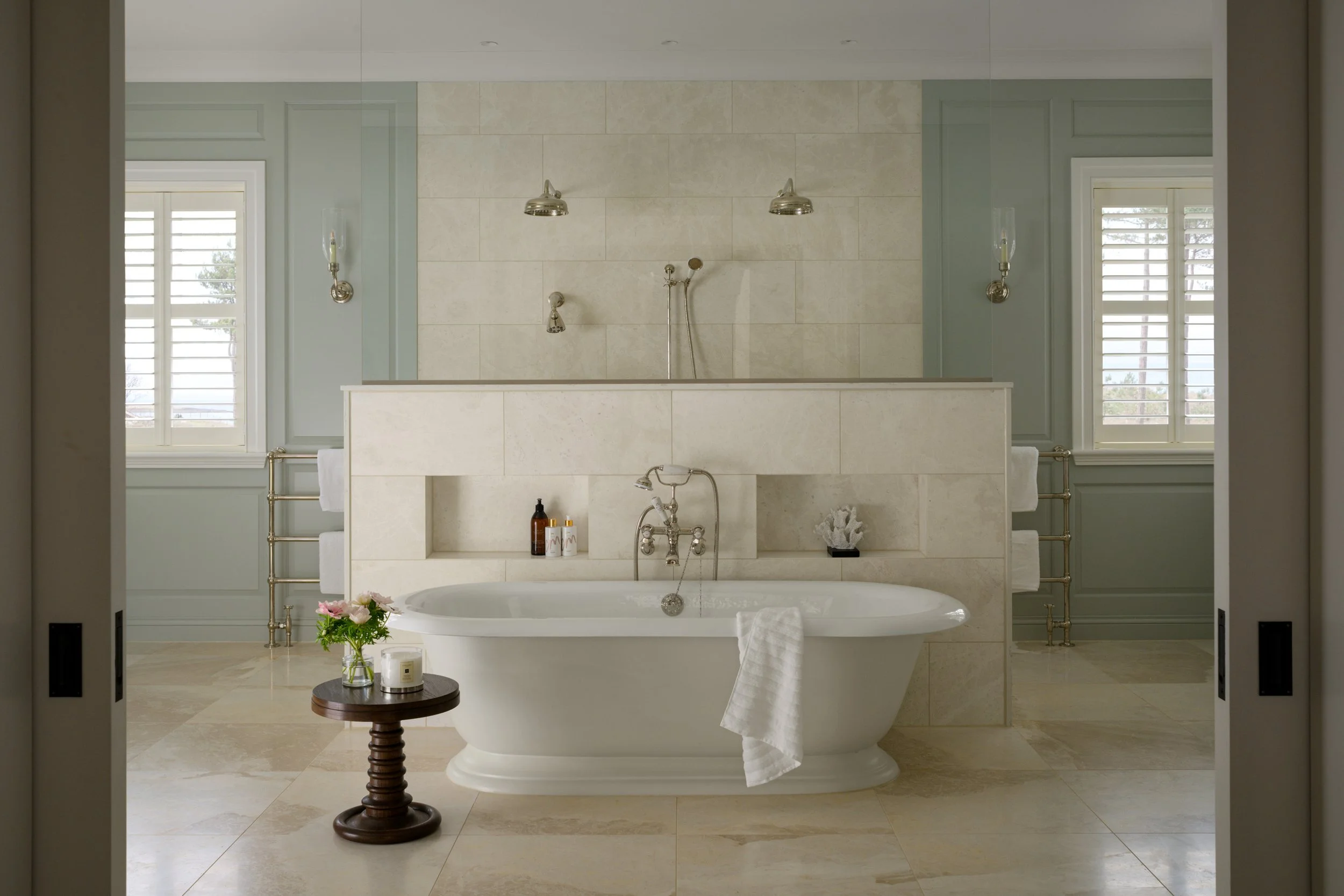 Elegant bathroom with a vintage bathtub, two windows with shutters, a small round table with flowers and candles, and soft neutral tones.