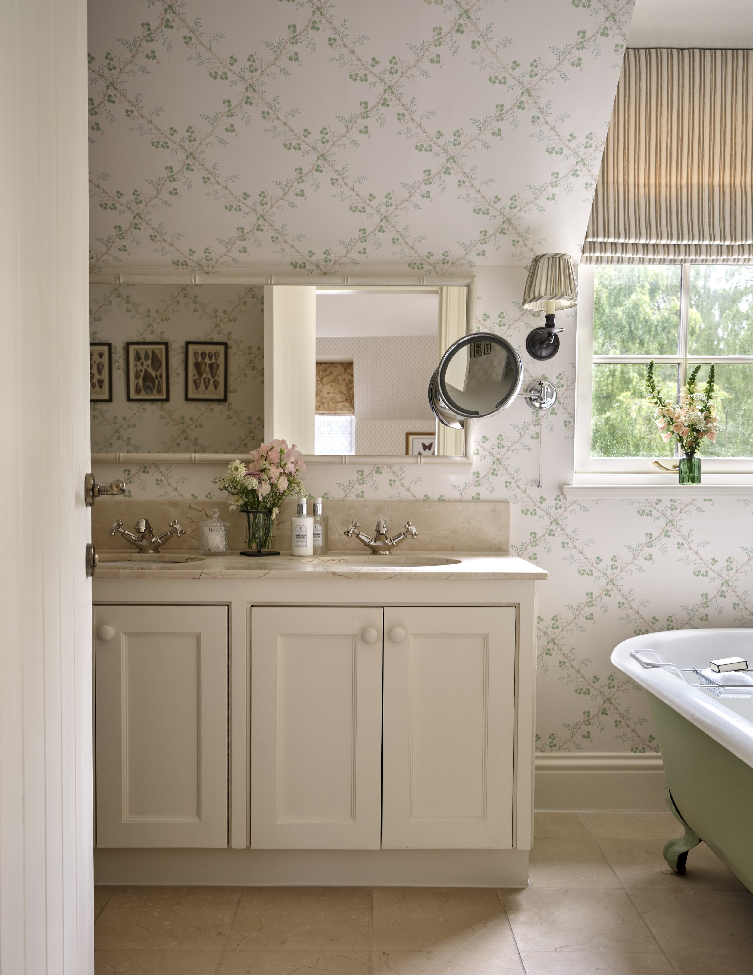 Bathroom with double vanity, framed mirror, wall-mounted magnifying mirror, small window with floral curtain, vase with flowers, bathtub, and floral wallpaper.