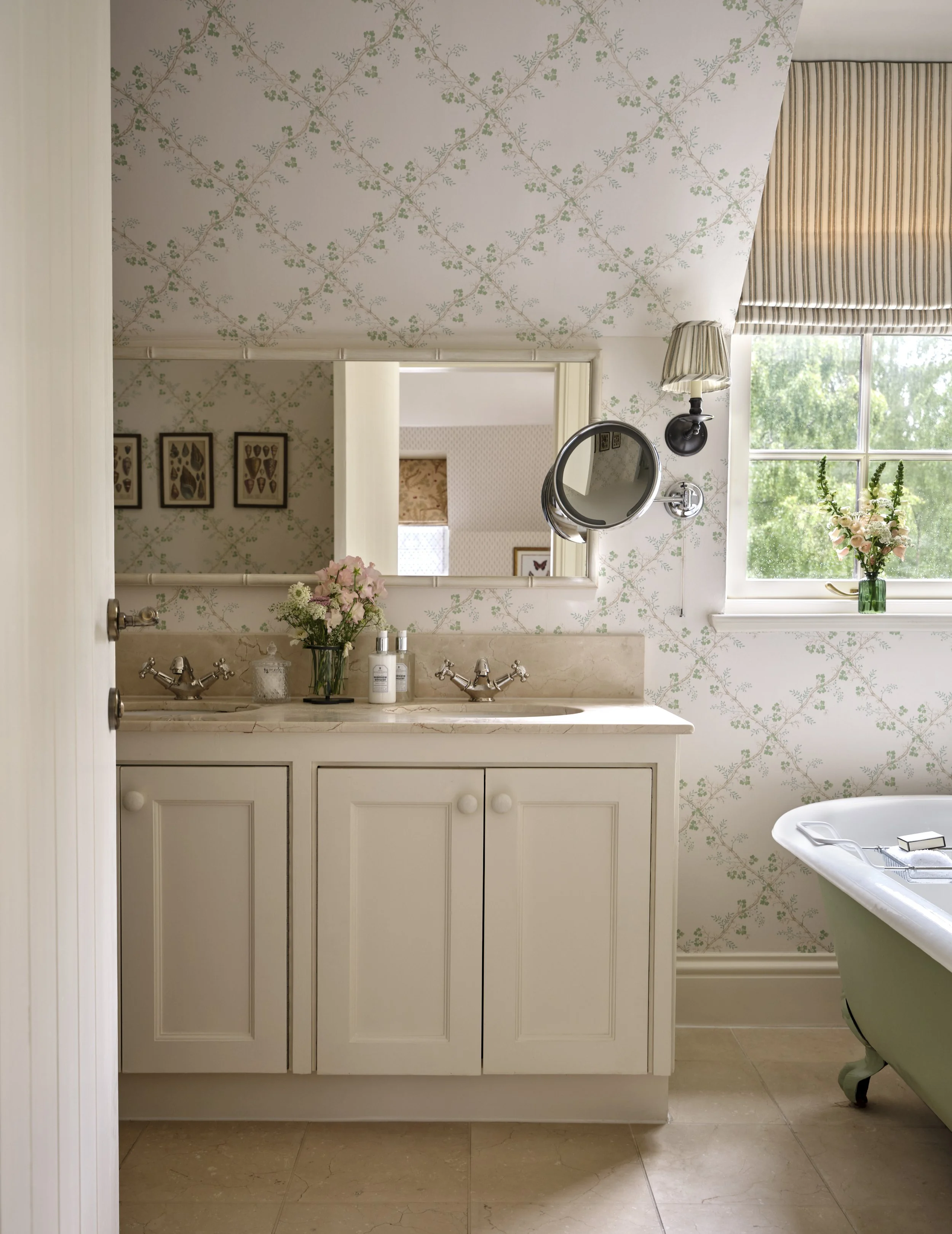 A vintage bathroom featuring a double sink vanity with pink marble countertop, floral wallpaper, a rectangular mirror, and a clawfoot bathtub with a green exterior near a window with a striped valance and a flower vase.