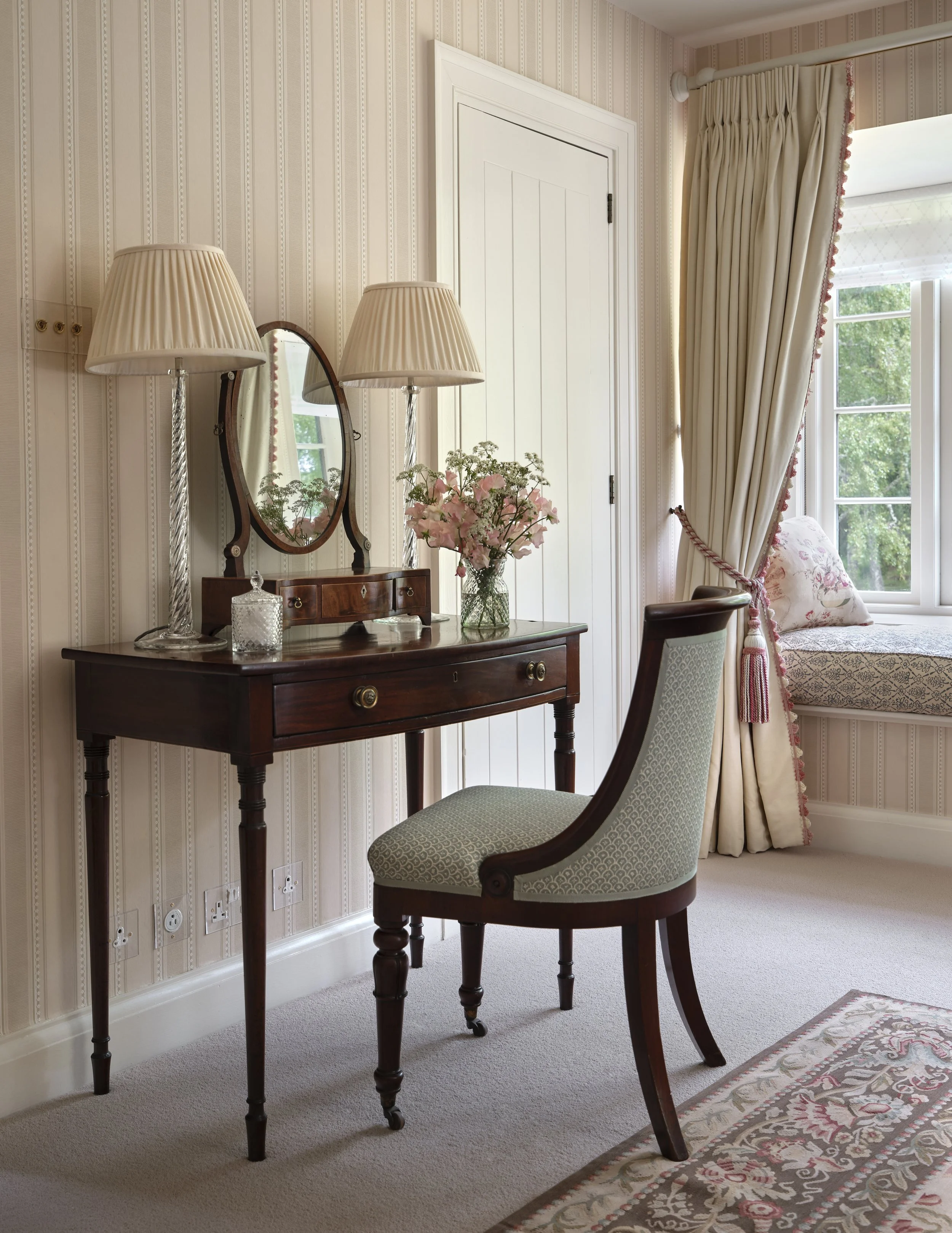 A vintage wooden vanity table with a large mirror, two table lamps, a glass vase with pink flowers, a beige upholstered chair, a window seat with floral cushion, heavy cream curtains, and a window showing greenery outside.
