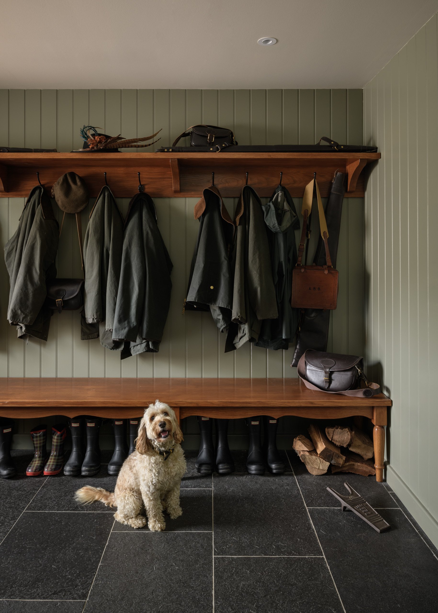 A mudroom with green paneled walls and black tile floor, featuring coat hooks with jackets, hats, and bags hanging, a wooden bench with boots underneath, and a small dog sitting on the floor.