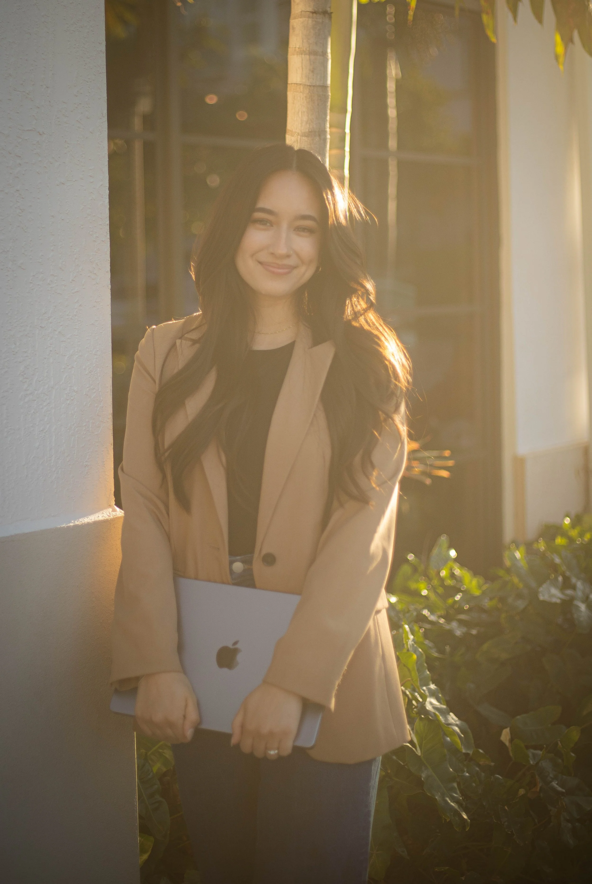 A woman with long dark hair holding a closed MacBook laptop, standing outdoors near a white wall, with sunlight shining behind her.