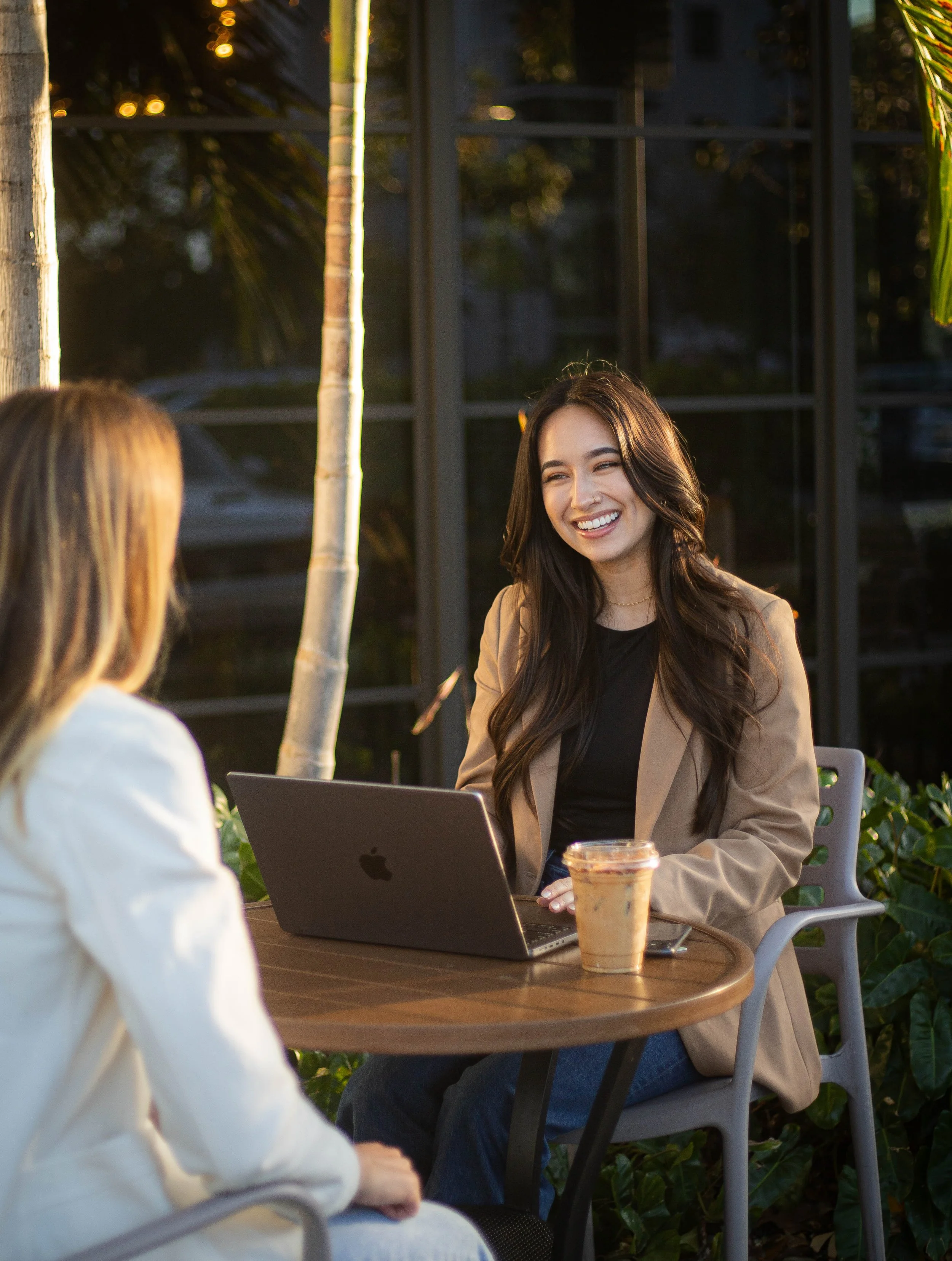 Two women having a conversation at an outdoor cafe, with one woman smiling, a laptop, and an iced coffee on the table.
