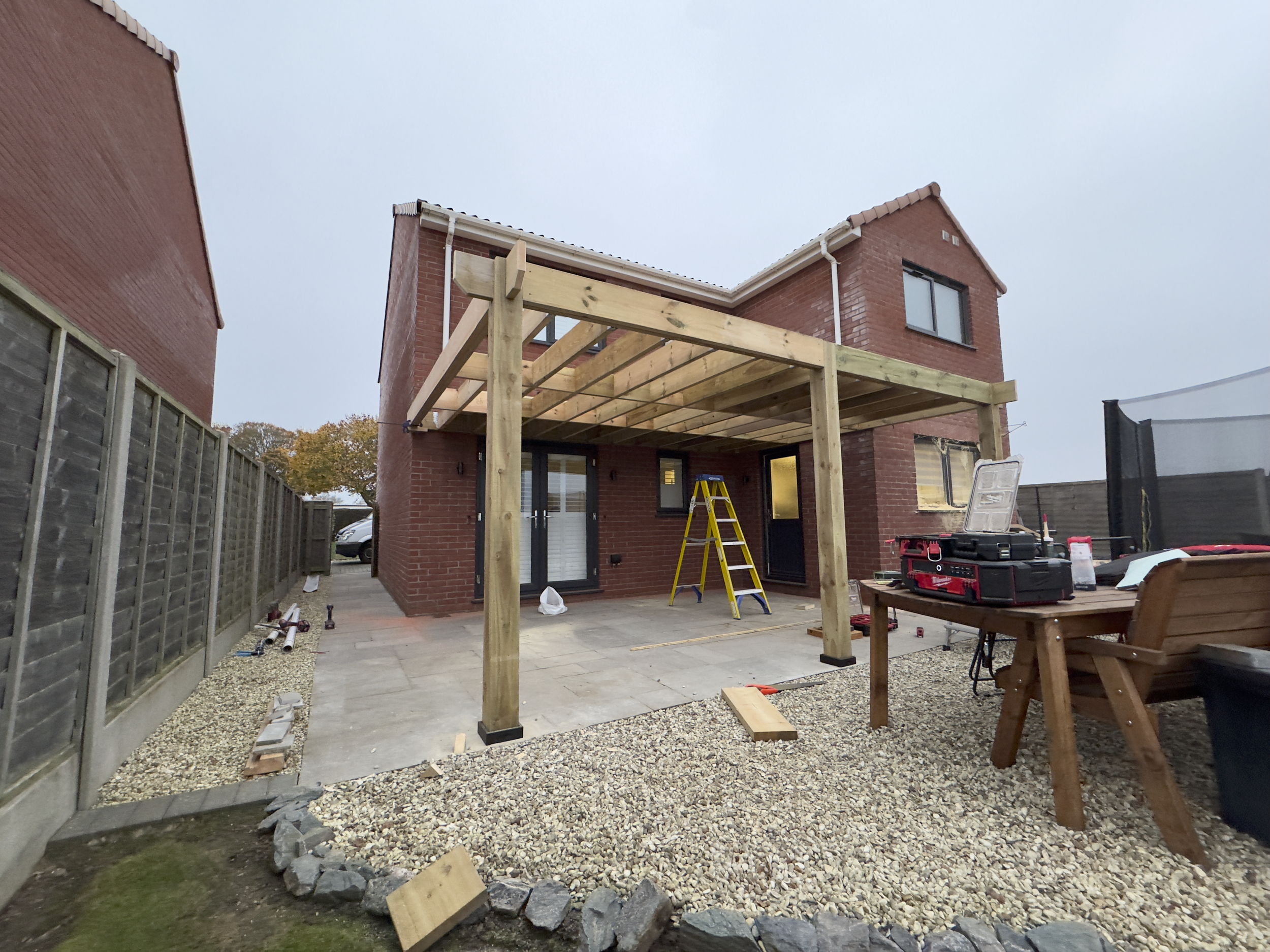 Backyard of a brick house under construction, with a wooden patio frame, a ladder, tools, and outdoor furniture.