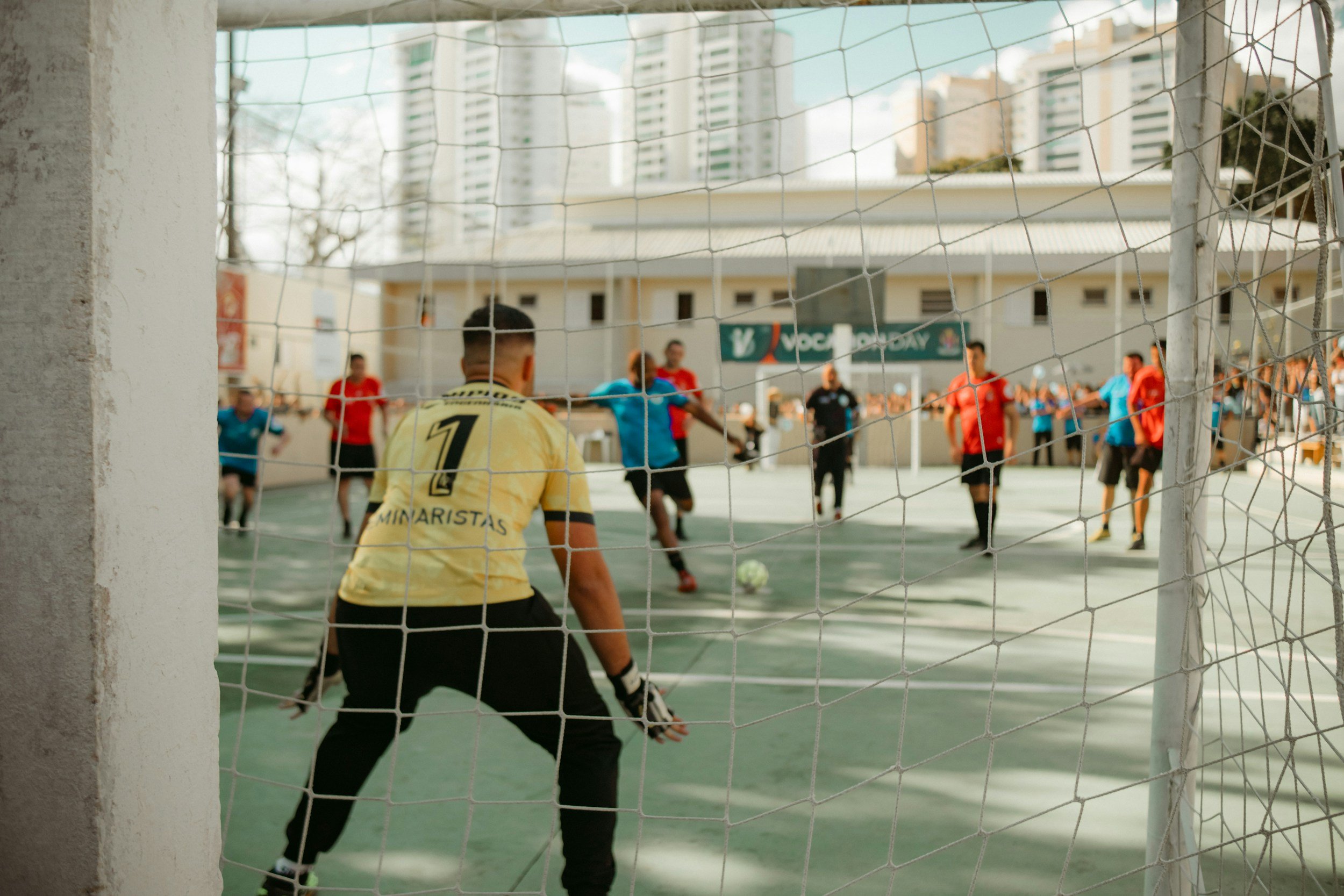 A soccer match on an outdoor court with players in blue, red, and yellow uniforms. The photo is taken from behind the goalkeeper wearing a yellow jersey with the number 1, standing in front of the goal net, with players actively pursuing the ball in the background.