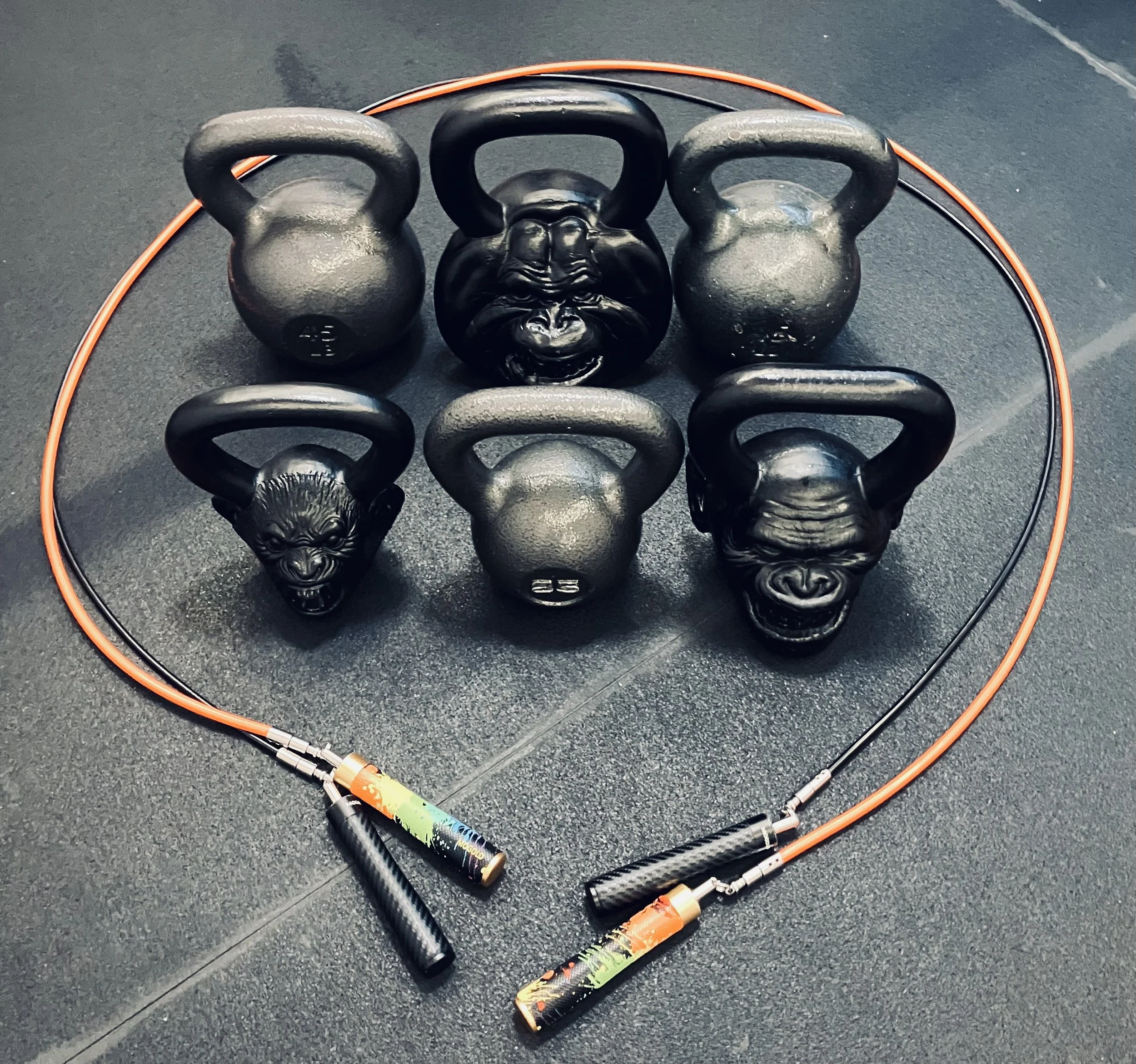 Six black kettlebells with various animal faces, arranged in two rows on a black gym floor. Two red and black jump ropes are coiled around them.