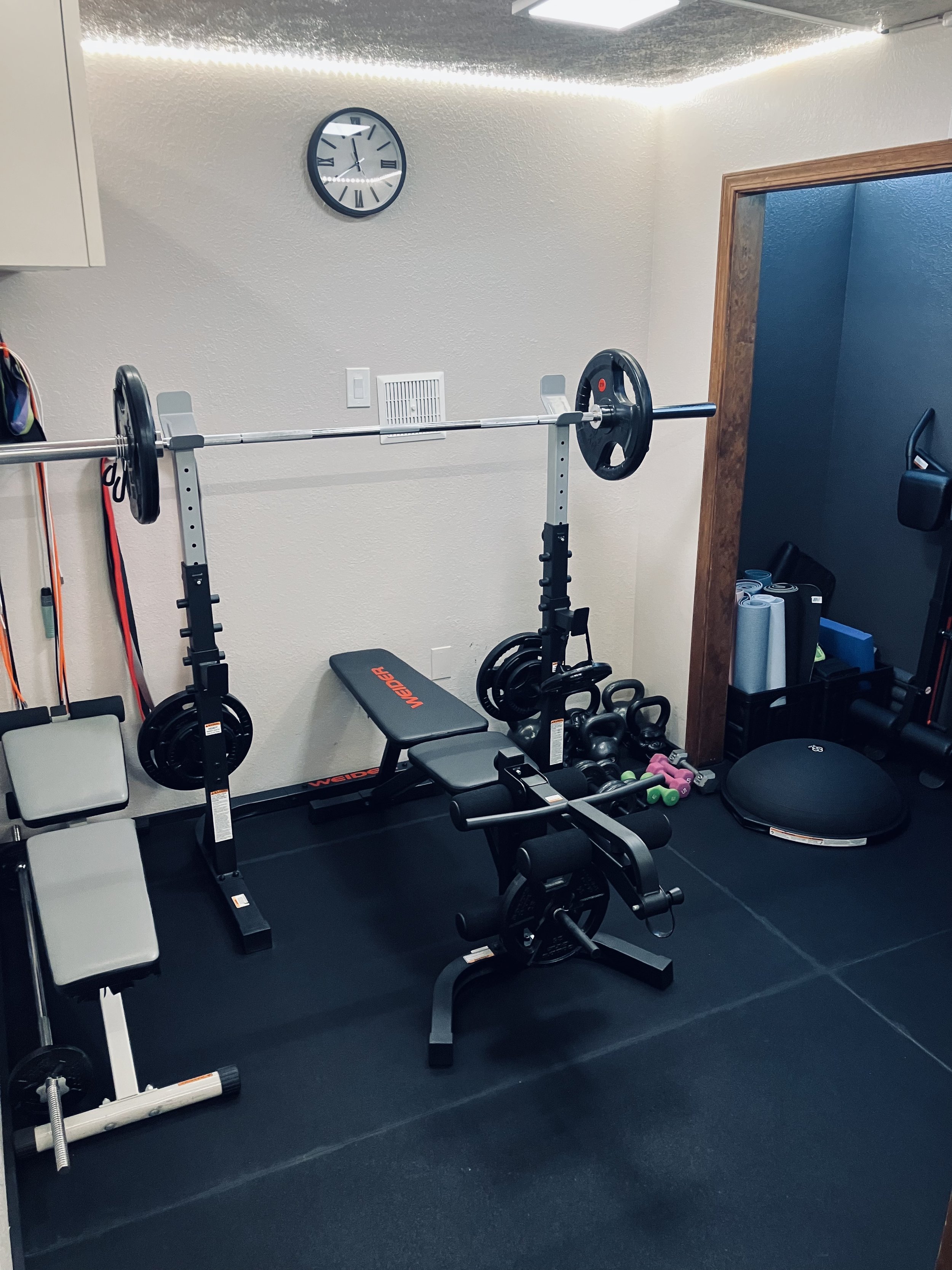 Home gym with weightlifting equipment, a clock on the wall, exercise mats, and dumbbells.