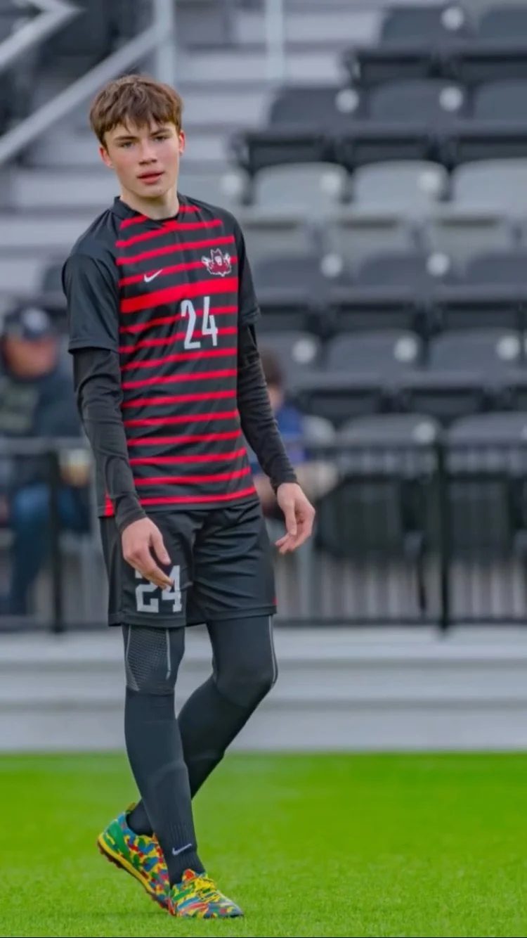 Young male soccer player standing on the field in a black and red striped jersey with the number 24, black shorts, black compression leggings, and colorful soccer cleats.