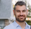 A man with dark hair and a beard smiling outdoors, wearing a checkered shirt with a garden in the background.
