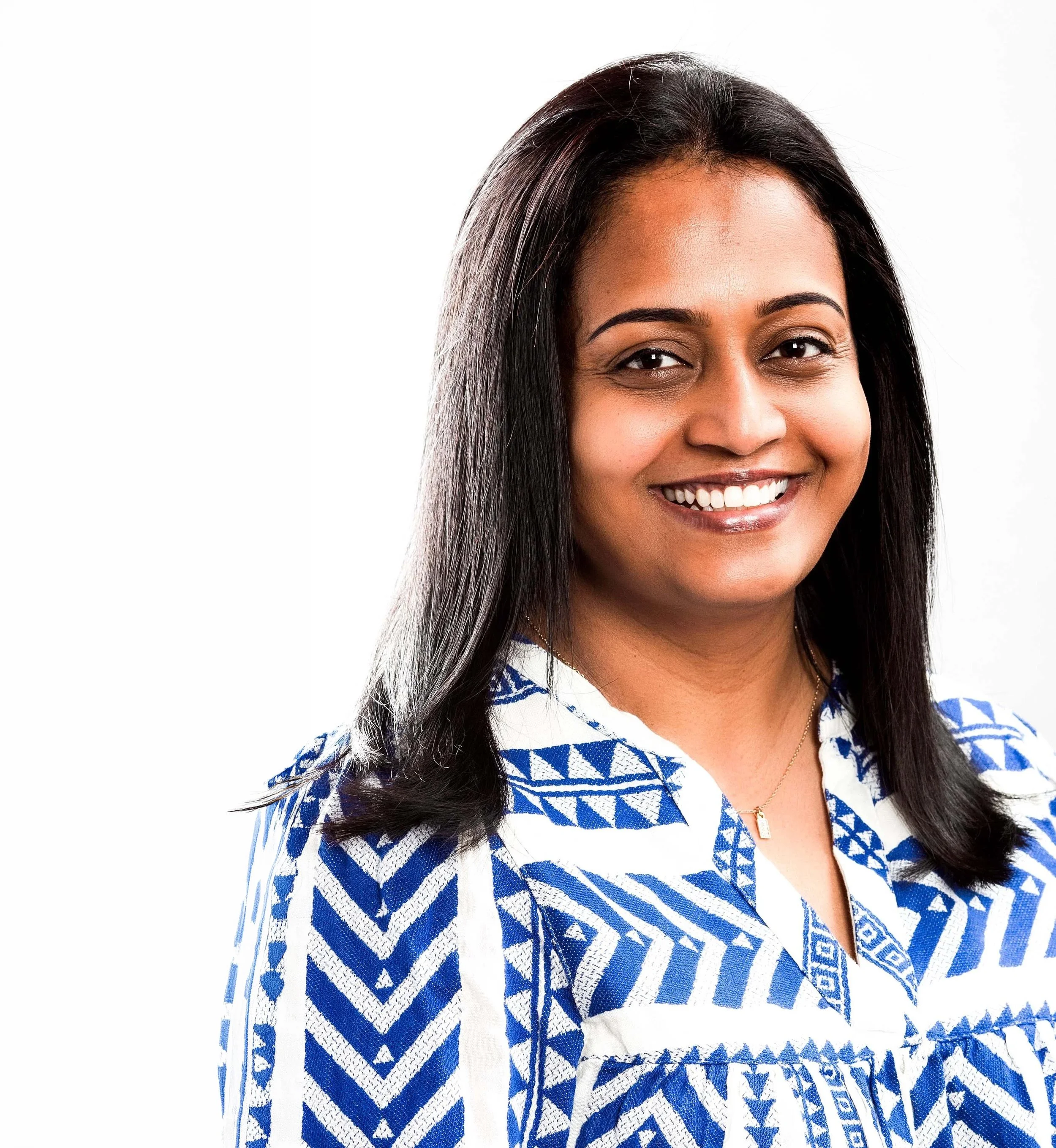 Close-up of a smiling woman with long black hair wearing a blue and white patterned outfit and a gold necklace, against a plain white background.