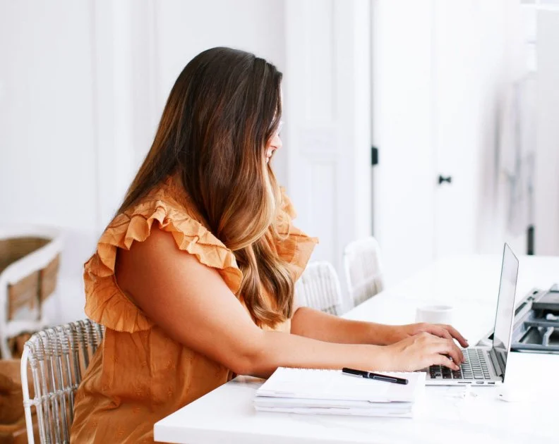 A woman with long brown hair sitting at a white table, working on a laptop, with notebooks and a pen in front of her, in a bright room.