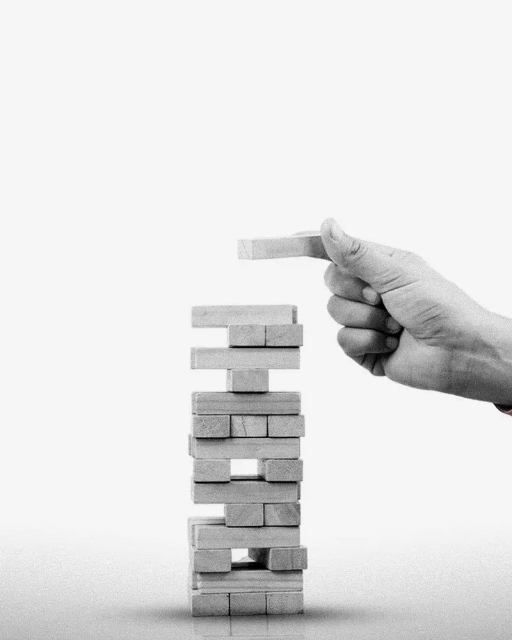 A hand in a glove removing a block from a vertical stack of wooden blocks, resembling a game of Jenga.