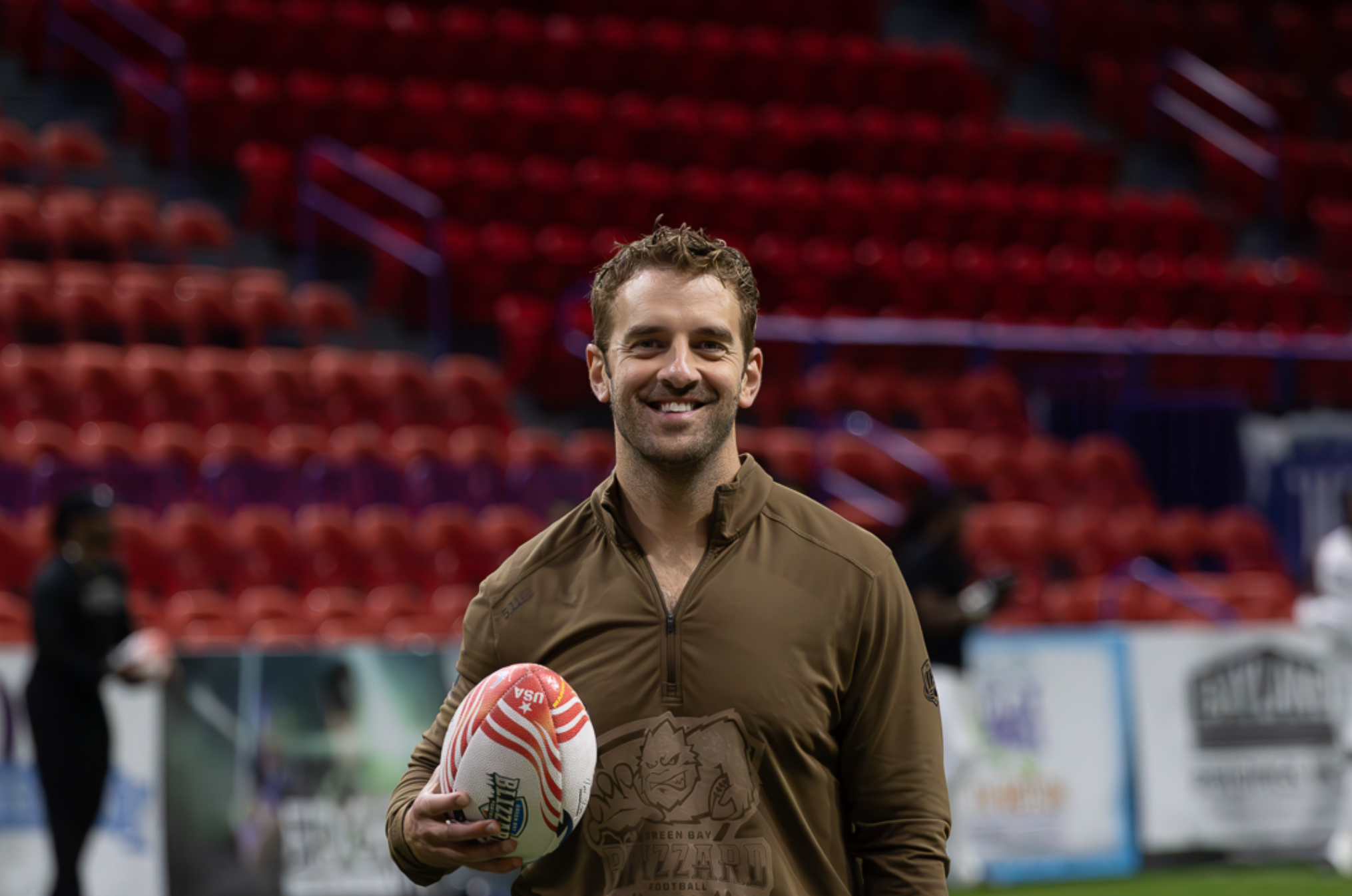 A man smiling and holding a rugby ball on a sports field with red stadium seats in the background.