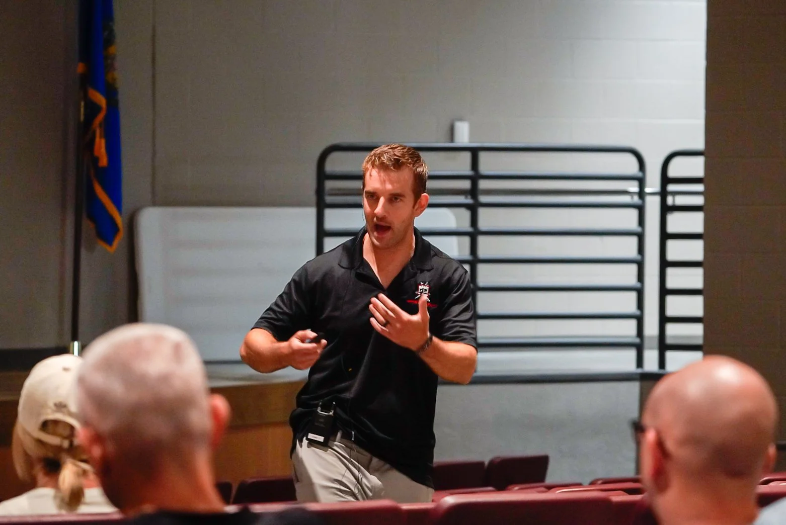 A man giving a presentation to an audience in a room, gesturing with his hand, with chairs and a flag in the background.