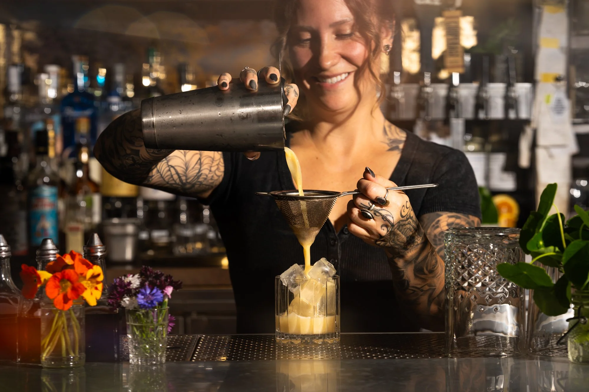 A female bartender with tattoos pours a yellow cocktail through a strainer into a glass with ice, in a bar with various bottles and plants around.