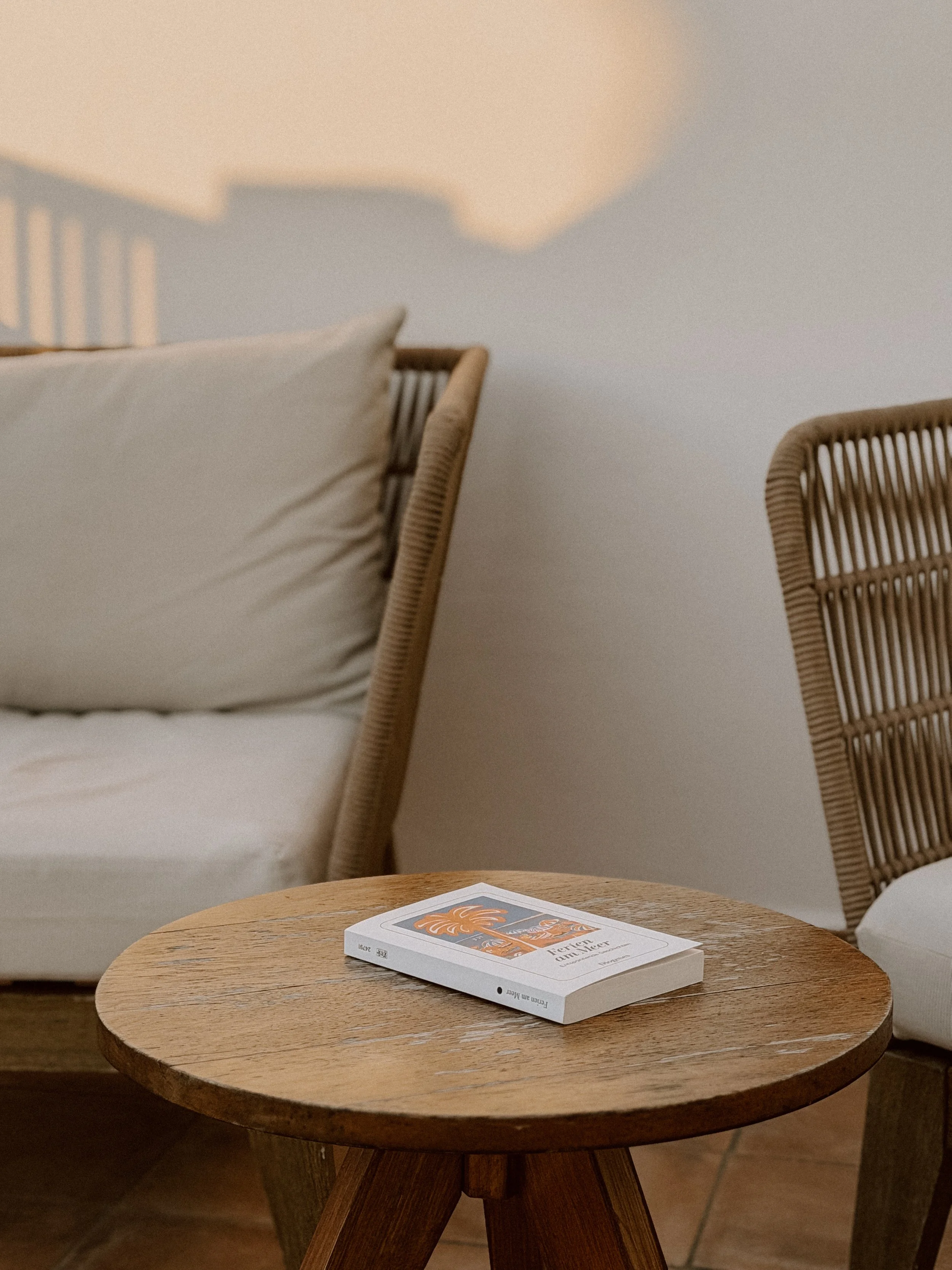 Ein runder Holztisch mit einem Buch darauf, umgeben von zwei Stühlen aus Rattan, auf einer Terrasse am Morgen.