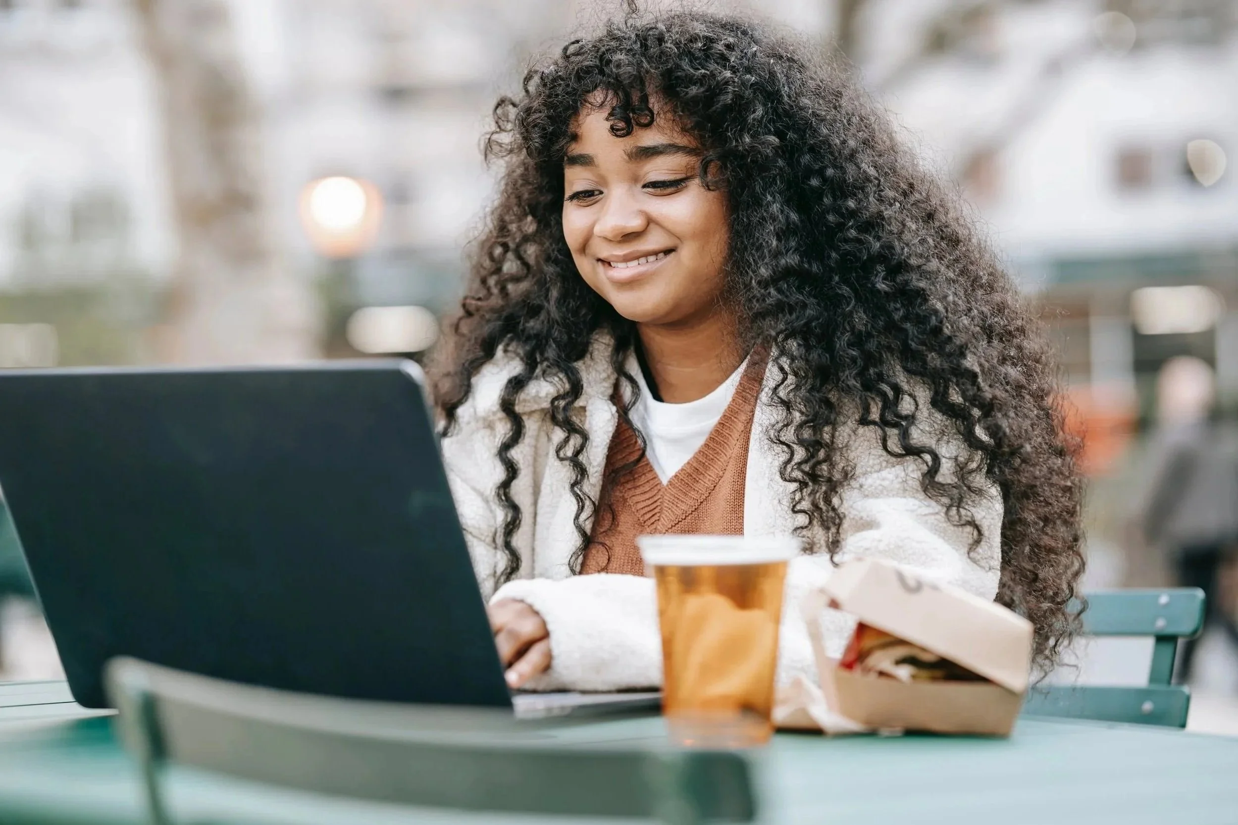 A woman with curly hair smiling at a laptop while sitting at a table outdoors, with a coffee cup and a newspaper in front of her.
