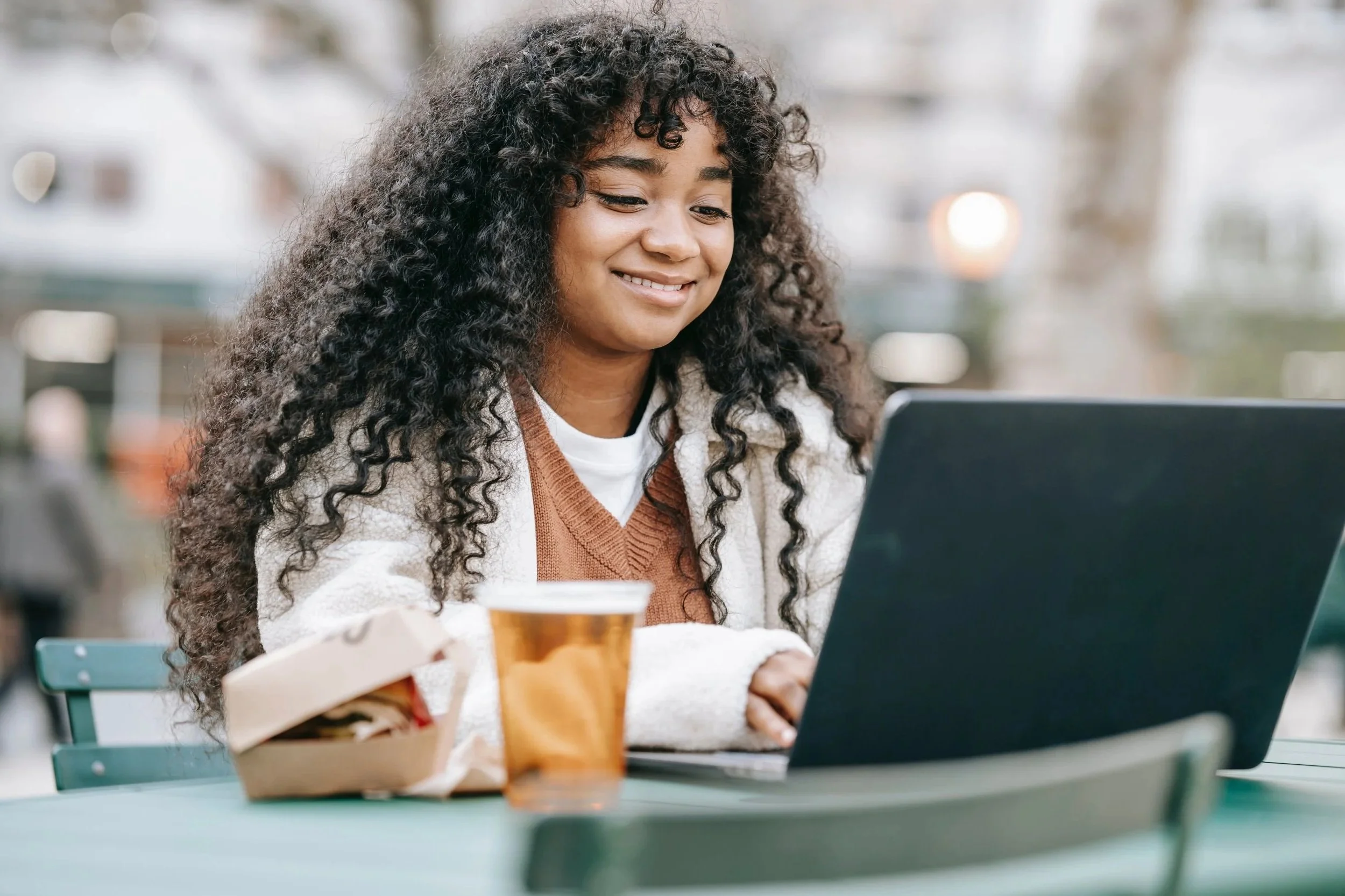 Young woman with curly hair smiling and looking at her laptop screen outdoors at a cafe table, with coffee and food nearby.
