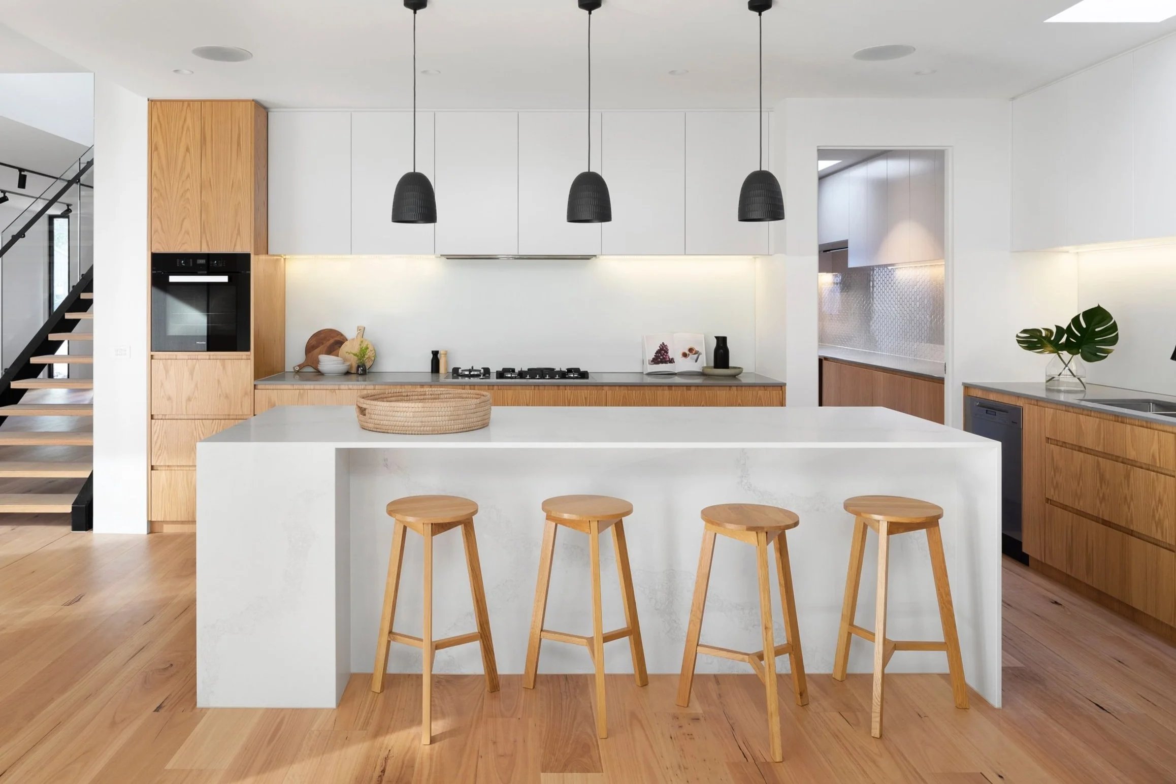 Modern kitchen with a white island, three wooden stools, hanging black pendant lights, wooden and white cabinetry, and a plant on the counter.