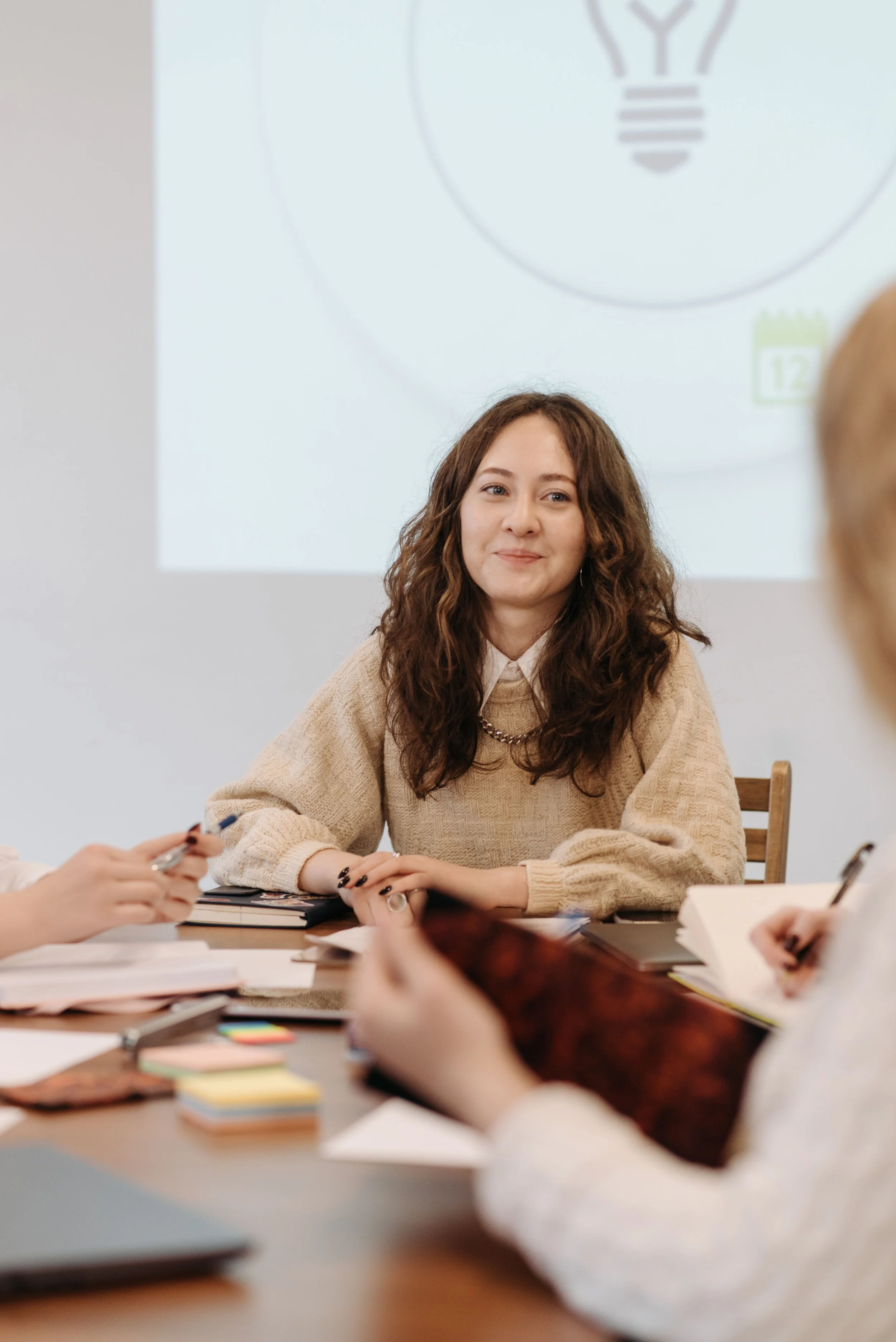 A young woman with wavy brown hair and wearing a beige sweater is seated at a table during a meeting. She is smiling and looking slightly to her right. Several people are around the table, some with notebooks, pens, and papers. The background features a whiteboard or presentation slide with a lightbulb illustration and a calendar icon.