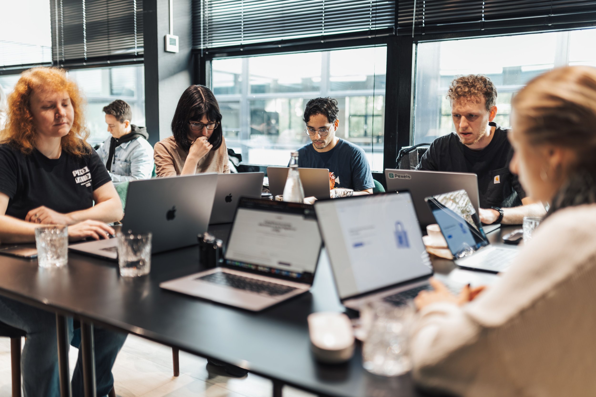 People sitting around a table working on laptops in a modern office space with large windows and natural light.