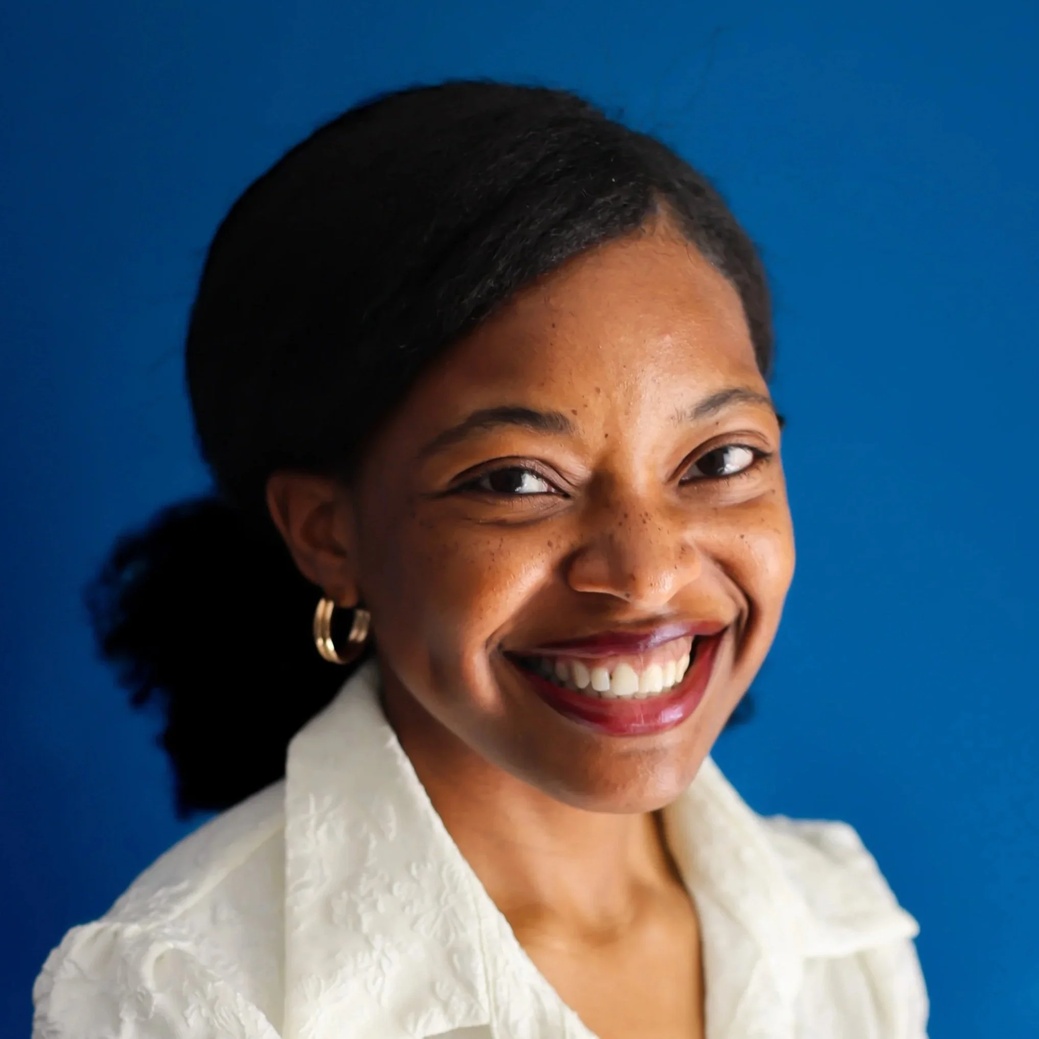Close-up of a woman smiling, wearing a white textured blazer and gold hoop earrings, against a blue background.