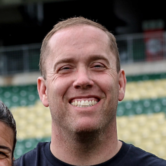 Close-up of a smiling man with short light hair in an outdoor sports stadium.