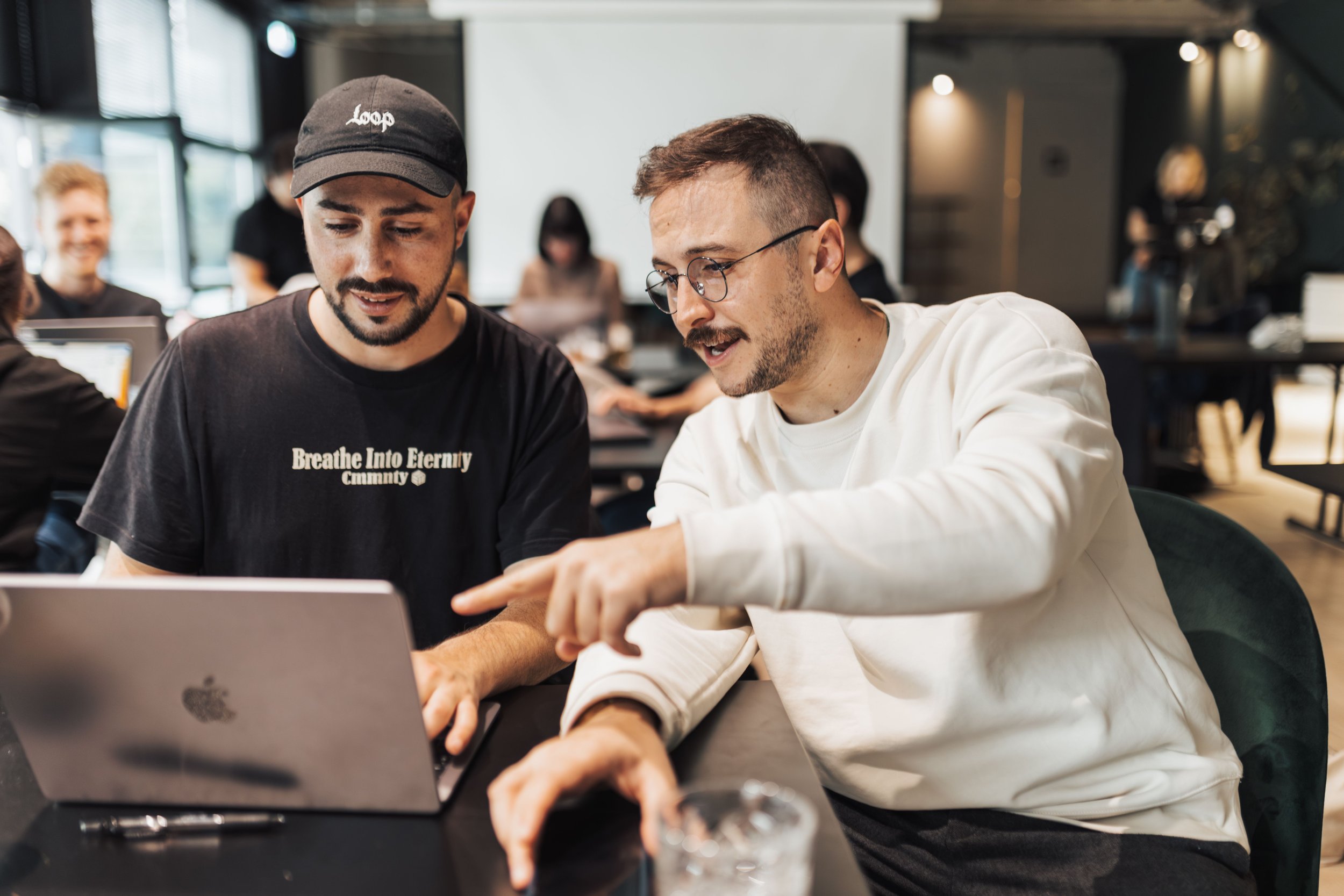 Two men are sitting at a table, looking at a laptop, in a busy modern cafe with other people in the background.