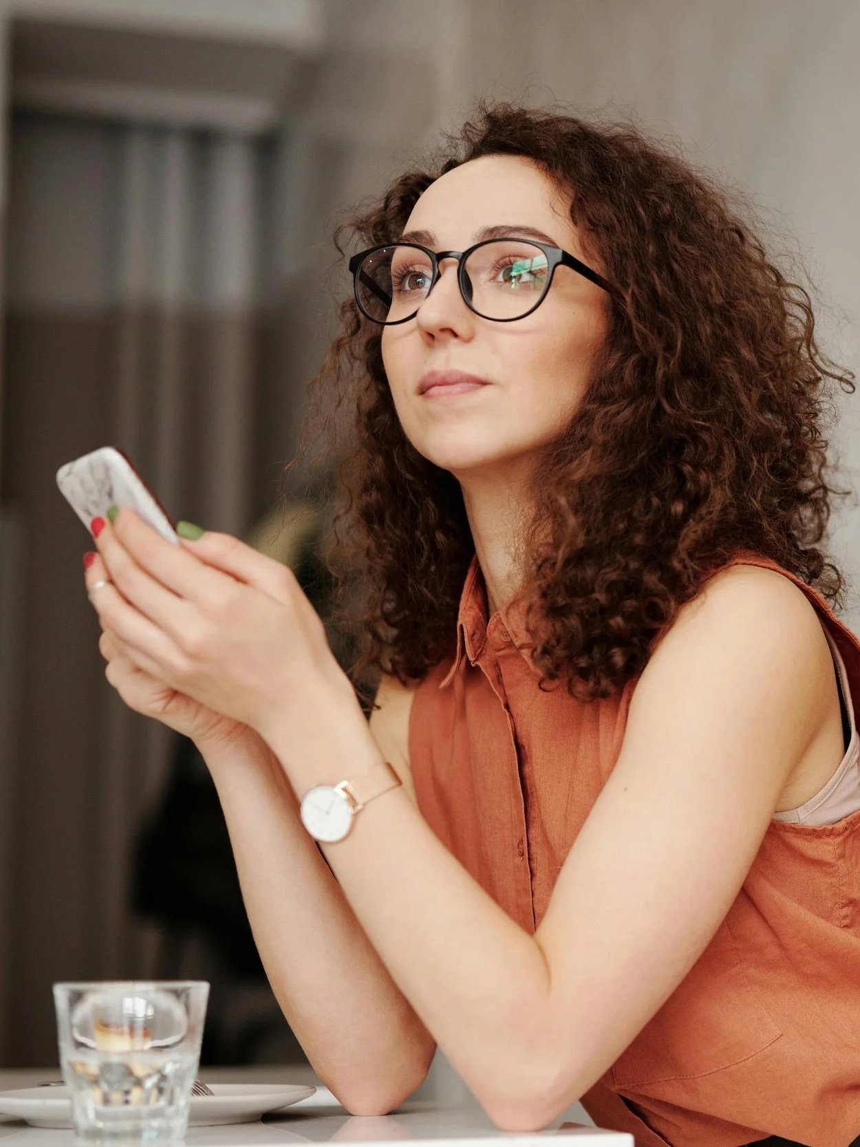 A woman with curly brown hair, glasses, and light skin sitting at a table, holding a smartphone with both hands, wearing a sleeveless orange top, looking thoughtful, with a glass of water and a plate in front of her.