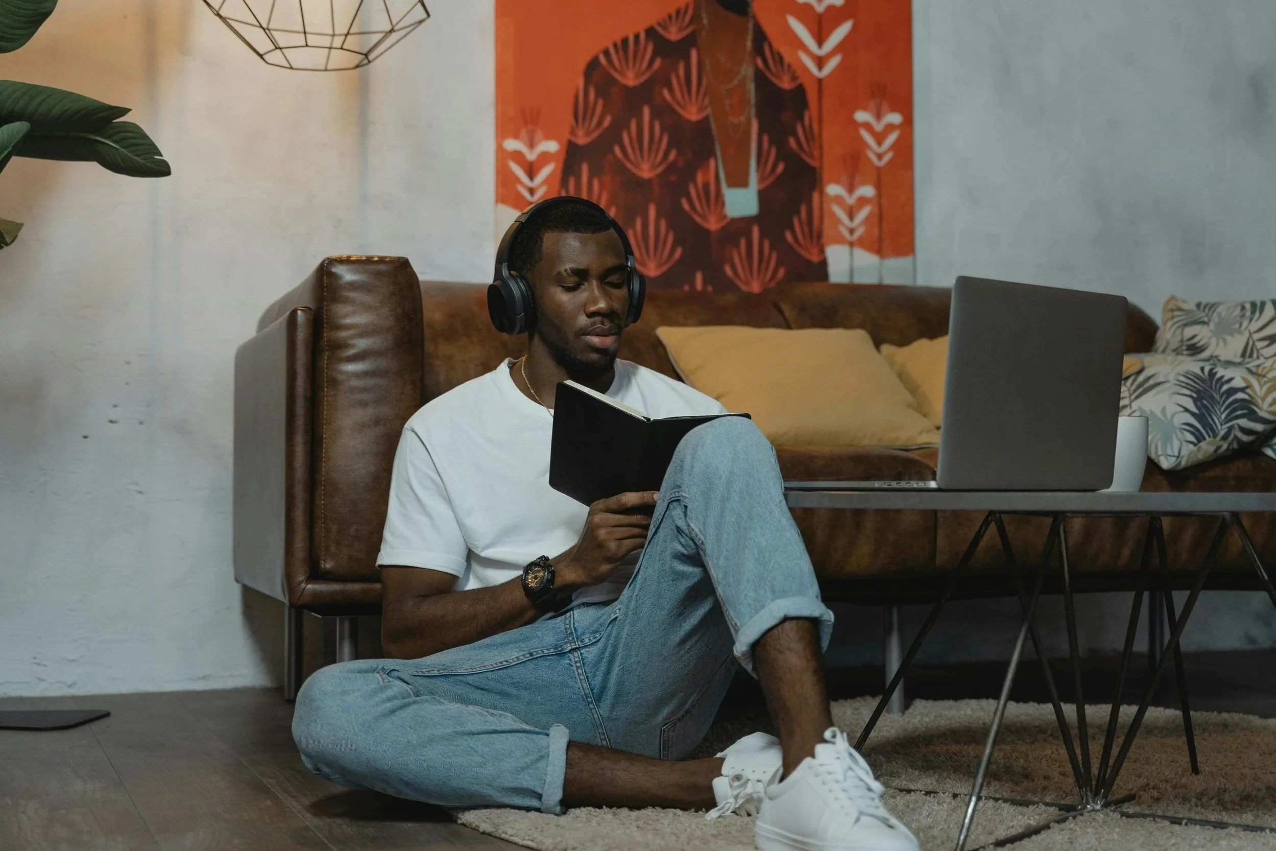 A young man with a dark complexion, wearing a white t-shirt and light blue jeans, sitting cross-legged on a brown leather armchair, listening to music with black headphones, and reading a black notebook. Behind him is a yellow pillow on a brown couch, and a colorful abstract artwork with red, black, and white patterns on the wall. A potted plant with large green leaves is visible on the left, and a decorative wireframe pendant light hangs from the ceiling.