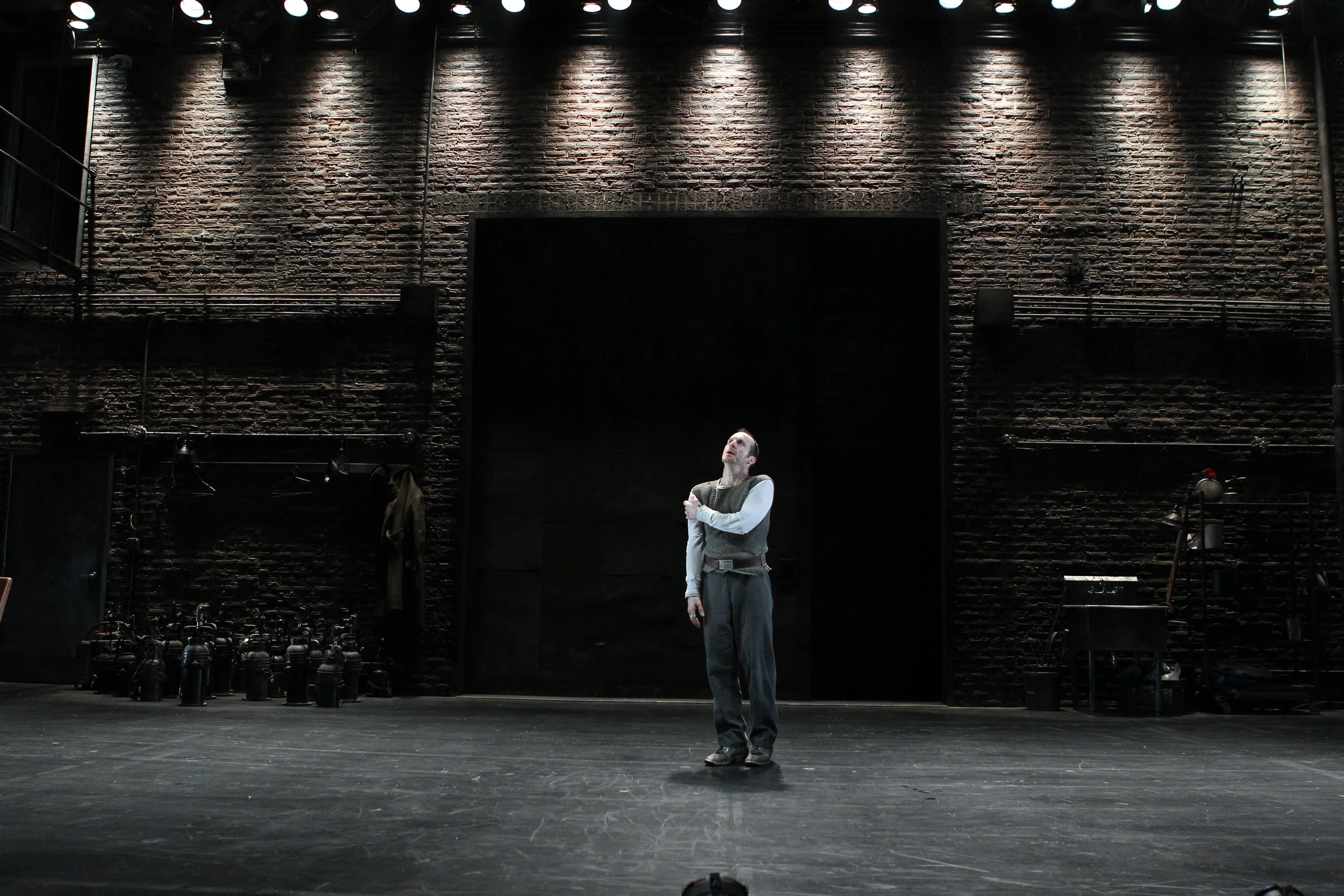 A person stands alone on a dark theater stage, lit by overhead stage lights, against a textured brick wall background. The stage has minimal props, including a coat hanging on the wall and a cluster of fire extinguishers on the left side.