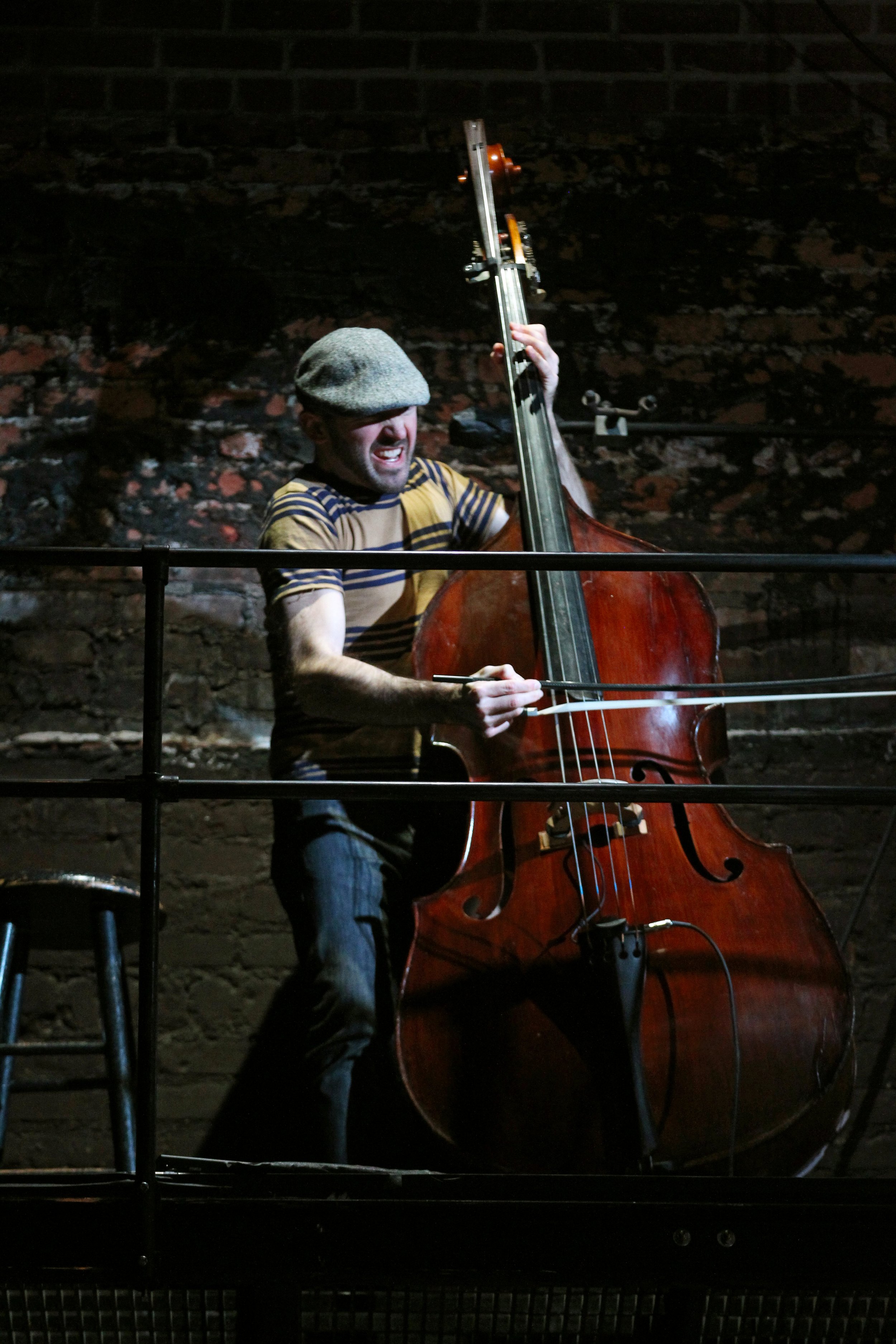 A man wearing a gray cap and a striped T-shirt playing a double bass on stage with dark, brick wall background.