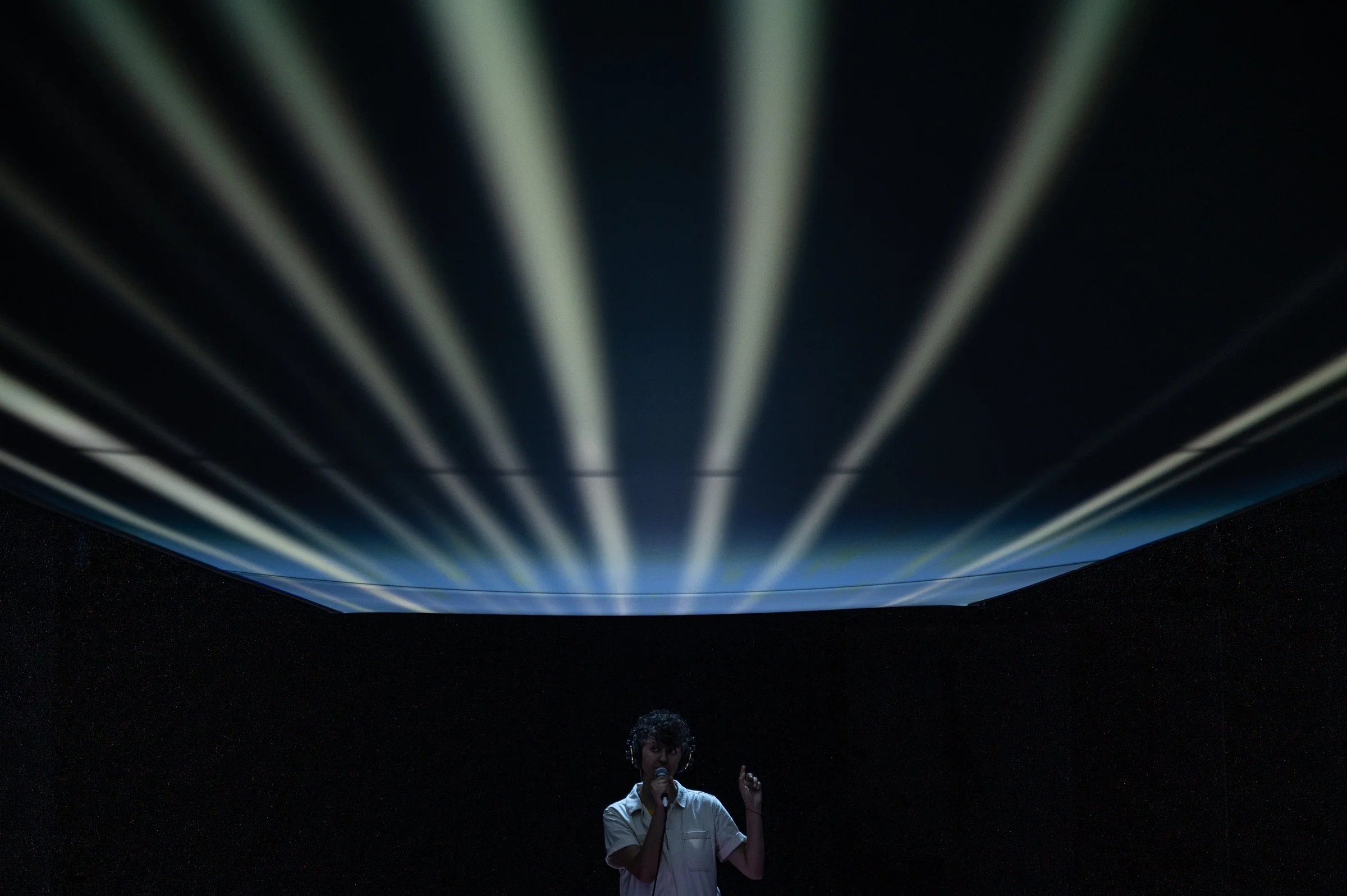 A person with headphones holding a microphone and pointing upwards, illuminated by a bright light and surrounded by dark space.