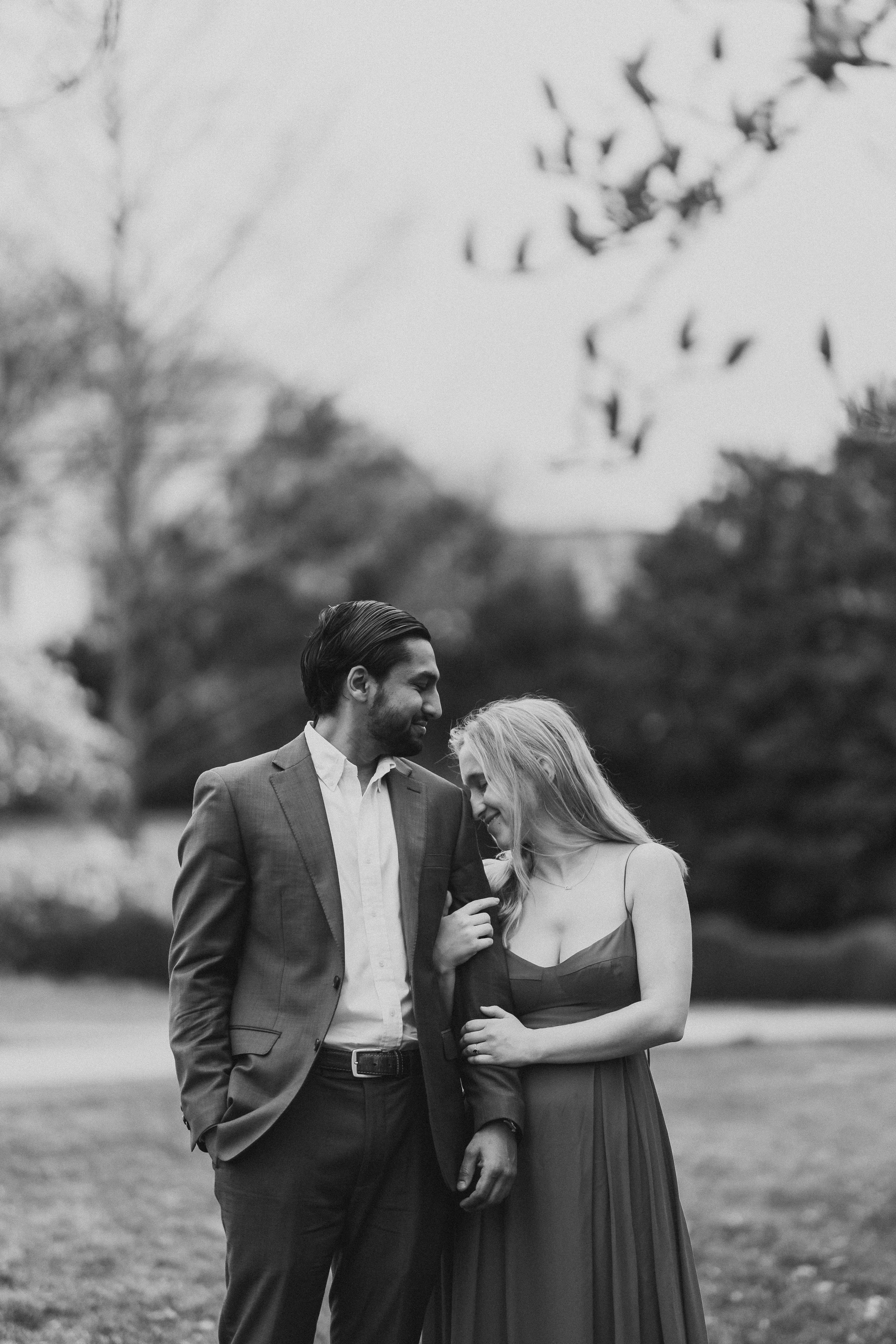 A black and white photo of a couple outdoors, with the man in a suit and the woman in a dress, smiling and sharing an intimate moment.