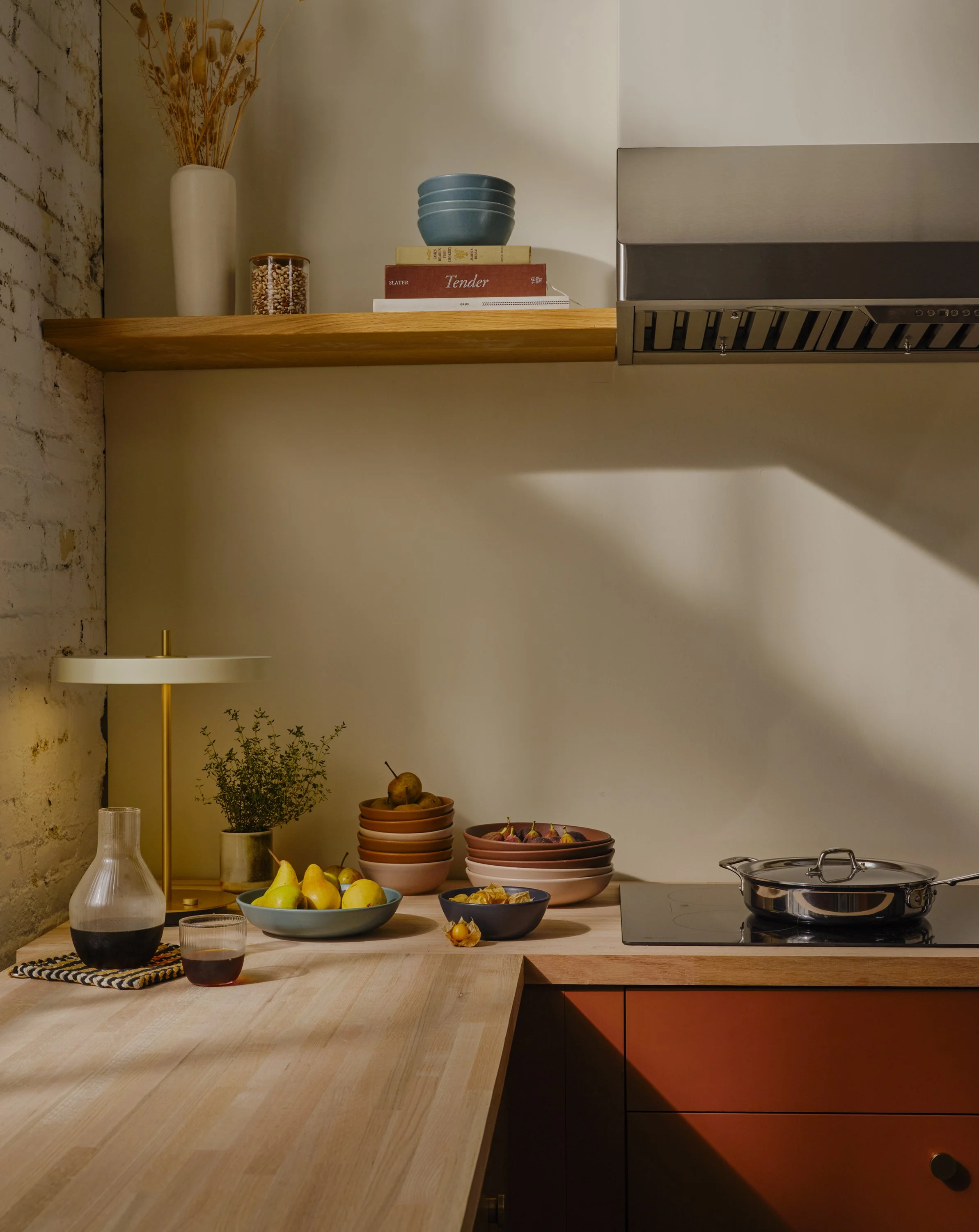 A kitchen countertop with a variety of bowls and fruits, a glass decanter with a dark liquid, a potted plant, a lamp, and a stove with a pot. There are stacked bowls and books on a wooden shelf above the counter.