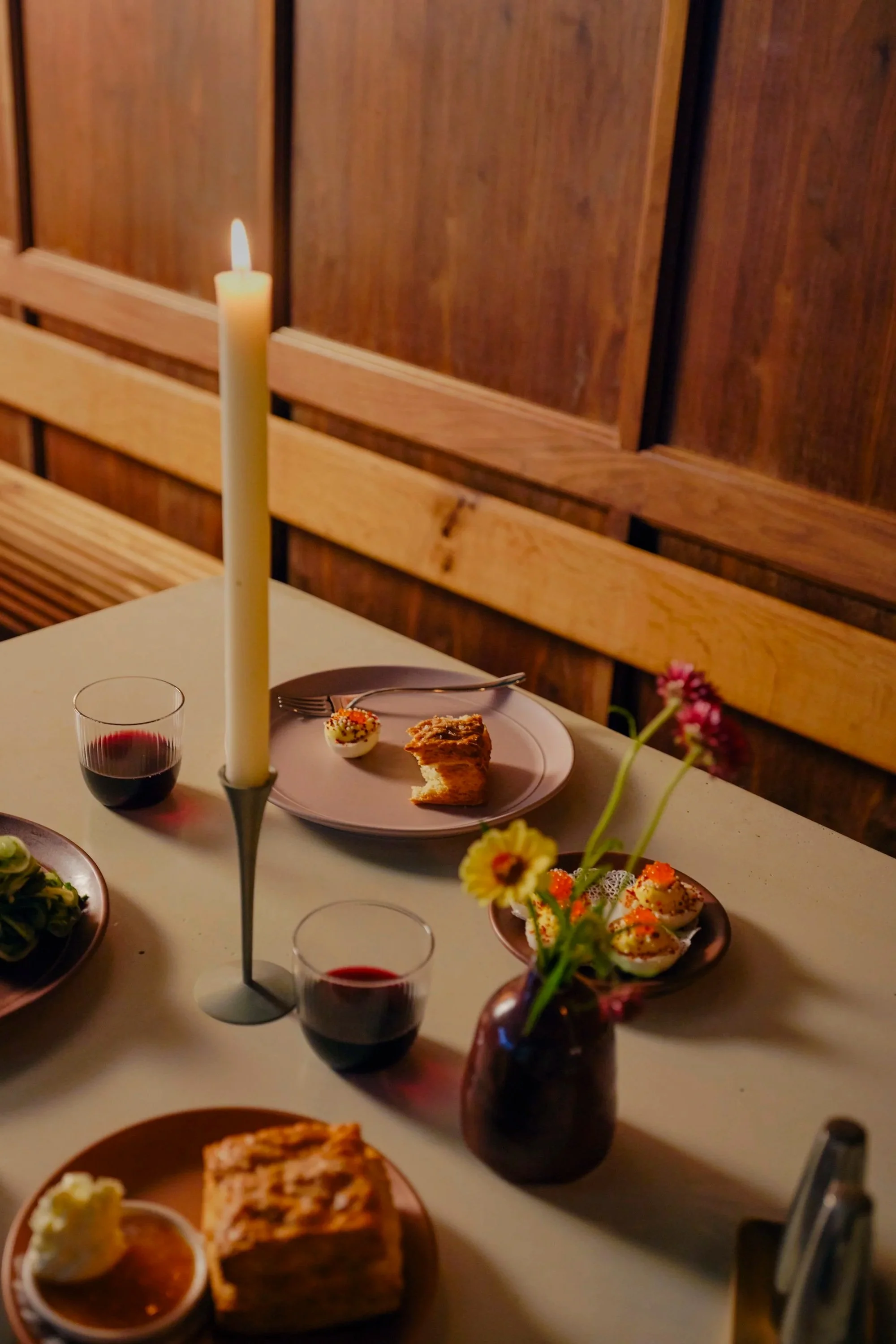 A dinner table with a white tablecloth, featuring a tall white candle in a silver holder, two glasses of red wine, and various small plates of food including cupcakes, bread, and salads. There is a wooden bench against a wooden wall in the background.