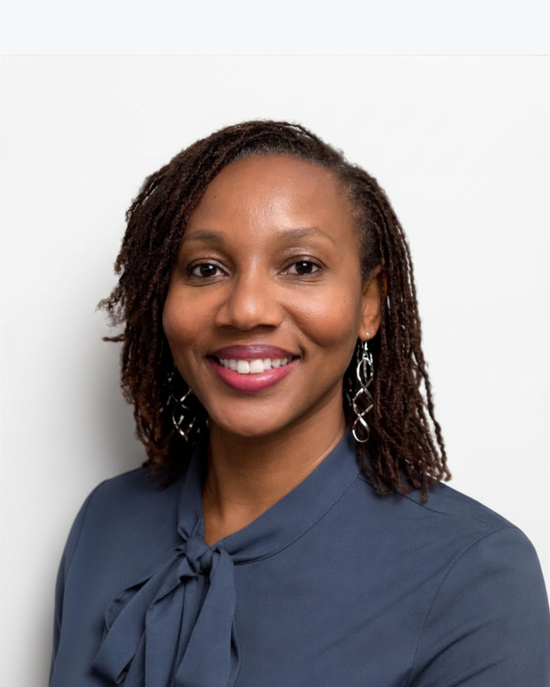 A professional woman with medium brown skin and shoulder-length dreadlocks, smiling, dressed in a navy blue top with a bow tie, wearing dangling silver earrings, against a plain white background.