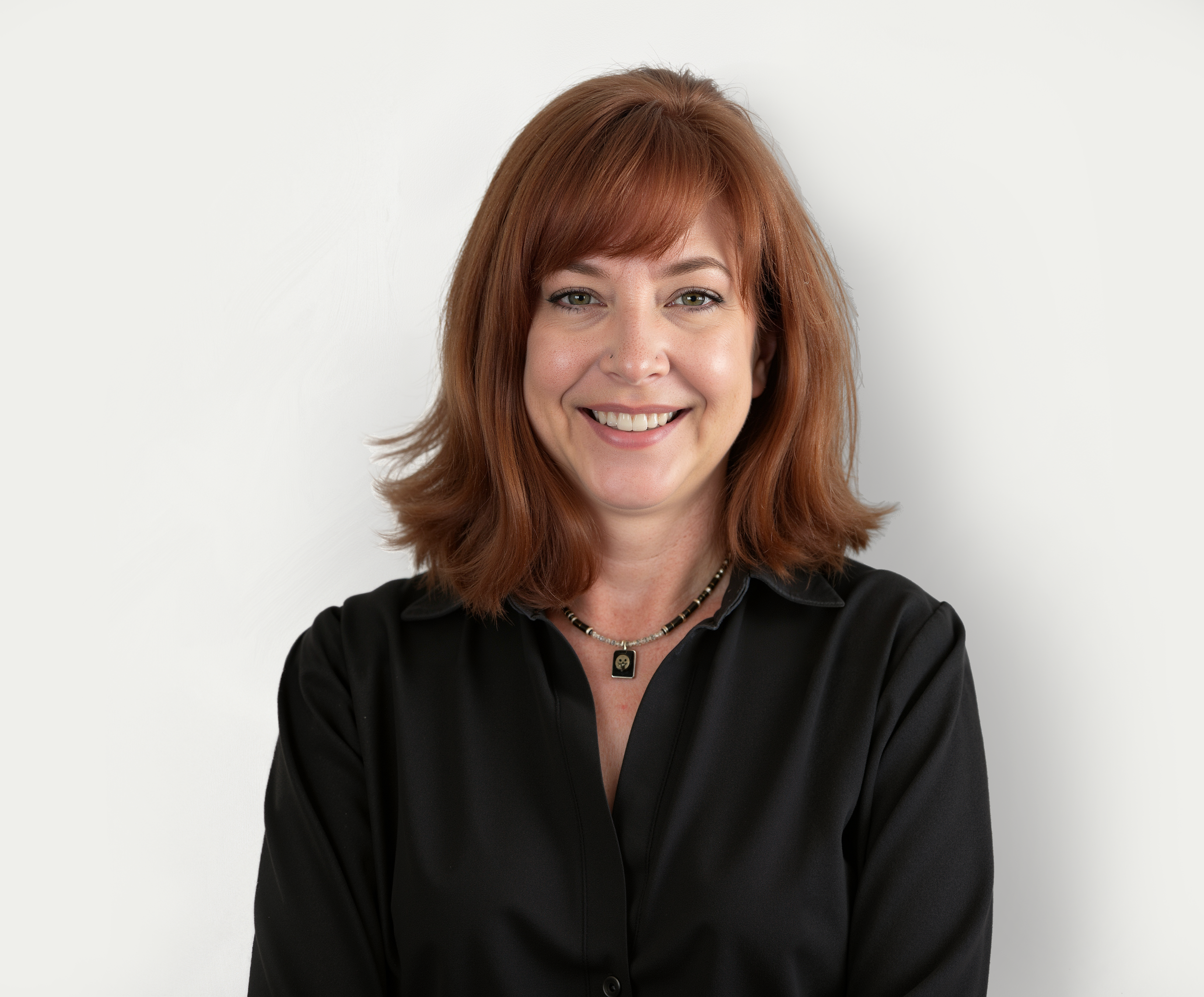 Smiling woman with red hair wearing a black shirt and a black necklace standing against a plain white wall.