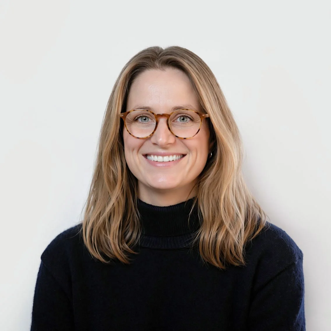 Portrait of a smiling woman with shoulder-length wavy blonde hair, wearing round tortoiseshell glasses and a black turtleneck sweater, against a plain light background.